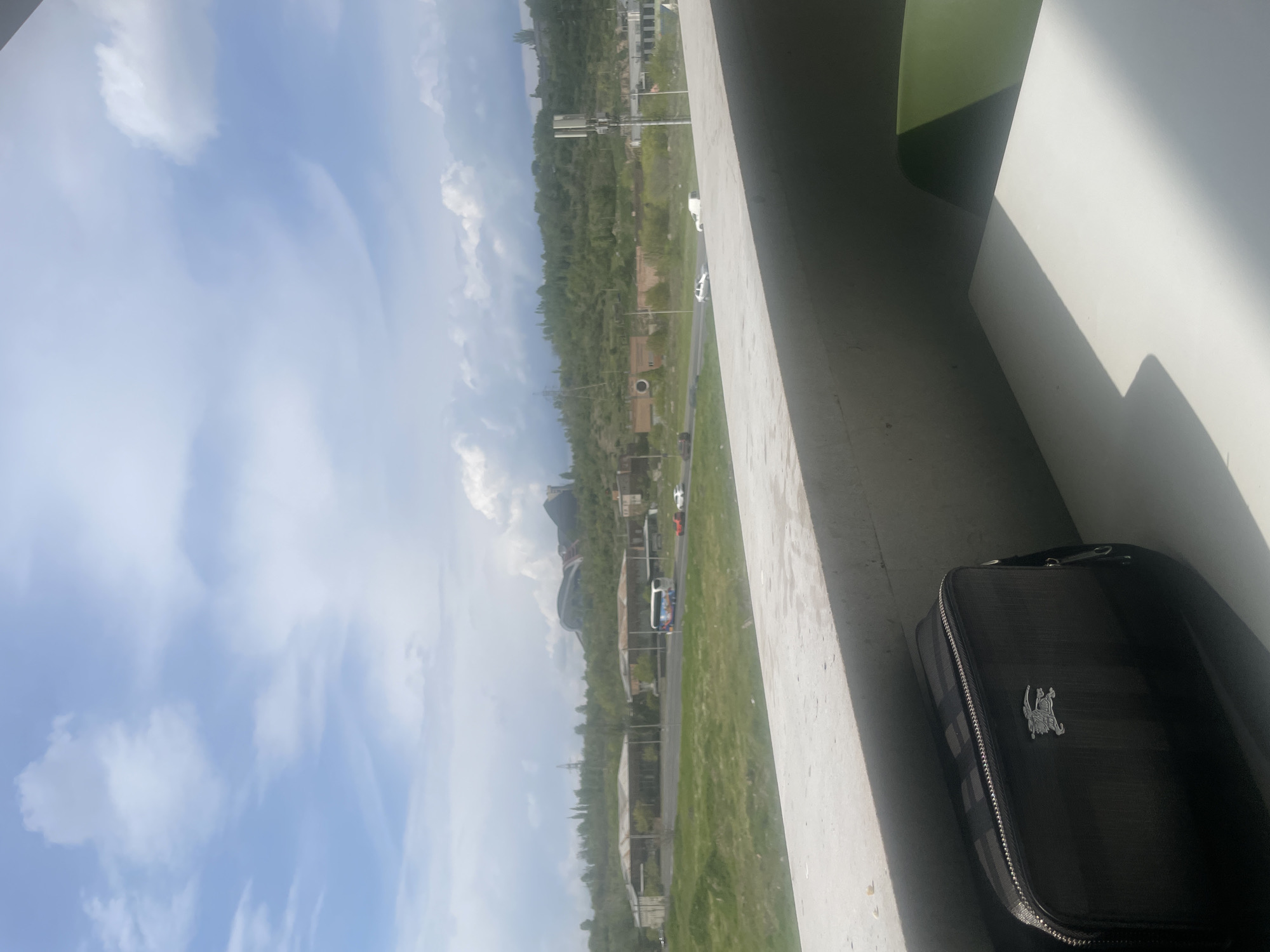 The image displays an outdoor scene captured from an elevated perspective, possibly a window or balcony. The sky occupies a significant portion of the view, showing a clear blue sky dotted with scattered white clouds, ranging from wispy to more substantial cumulus formations.
Below the sky, a verdant landscape extends into the distance, covered extensively with green trees and grassy terrain. A road or highway winds through this landscape, with several vehicles visible, including a white car, a light-colored bus, and a red car further down the road. In the mid-ground, various structures can be seen, notably a building with a distinctive dark, curved roof. Other less distinct buildings and utility poles are also present across the panorama.
In the foreground, a prominent concrete ledge or wall runs along the bottom and right side of the image, from which the view is taken. Bright sunlight casts strong, angular shadows on this concrete surface and on an upper right-hand corner, where a segment of a green wall or panel is visible. Resting on the concrete ledge in the lower right is a dark, patterned bag or case. This bag features a metallic emblem depicting a person on horseback, which is the recognizable logo for U.S. Polo Assn.
The strong shadows indicate that the time of day is likely mid-day or afternoon, under clear and sunny weather conditions. There is no specific event taking place, just a static view of the urban periphery with regular traffic. The location context provided for this scene is Yerevan, Armenia. No legible text is visible apart from the brand logo on the bag.