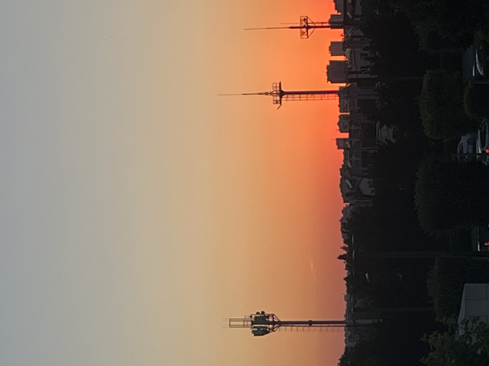 This is a vertical, full-frame photograph taken at sunset in Krasnodar, Russia. The sky displays a gradient of colors, transitioning from a pale gray-blue on the left to a vibrant orange-red on the right, indicating the sun is low on the horizon.
In the foreground, a thin, tall tower with a horizontal antenna structure at its top is silhouetted against the colorful sky. Further to the right, another similar tower is visible, also silhouetted. Beyond these towers, a cityscape is depicted, characterized by several multi-story buildings with distinct architectural features, partially obscured by dark silhouettes of trees. Cars are also visible in the right portion of the image, suggesting a residential or urban area.
The overall scene evokes a sense of calm and the end of the day. The light conditions are soft, typical of twilight. There is no visible text in the image. The composition and the presence of towers suggest the possibility of a communication or broadcasting hub within the city.