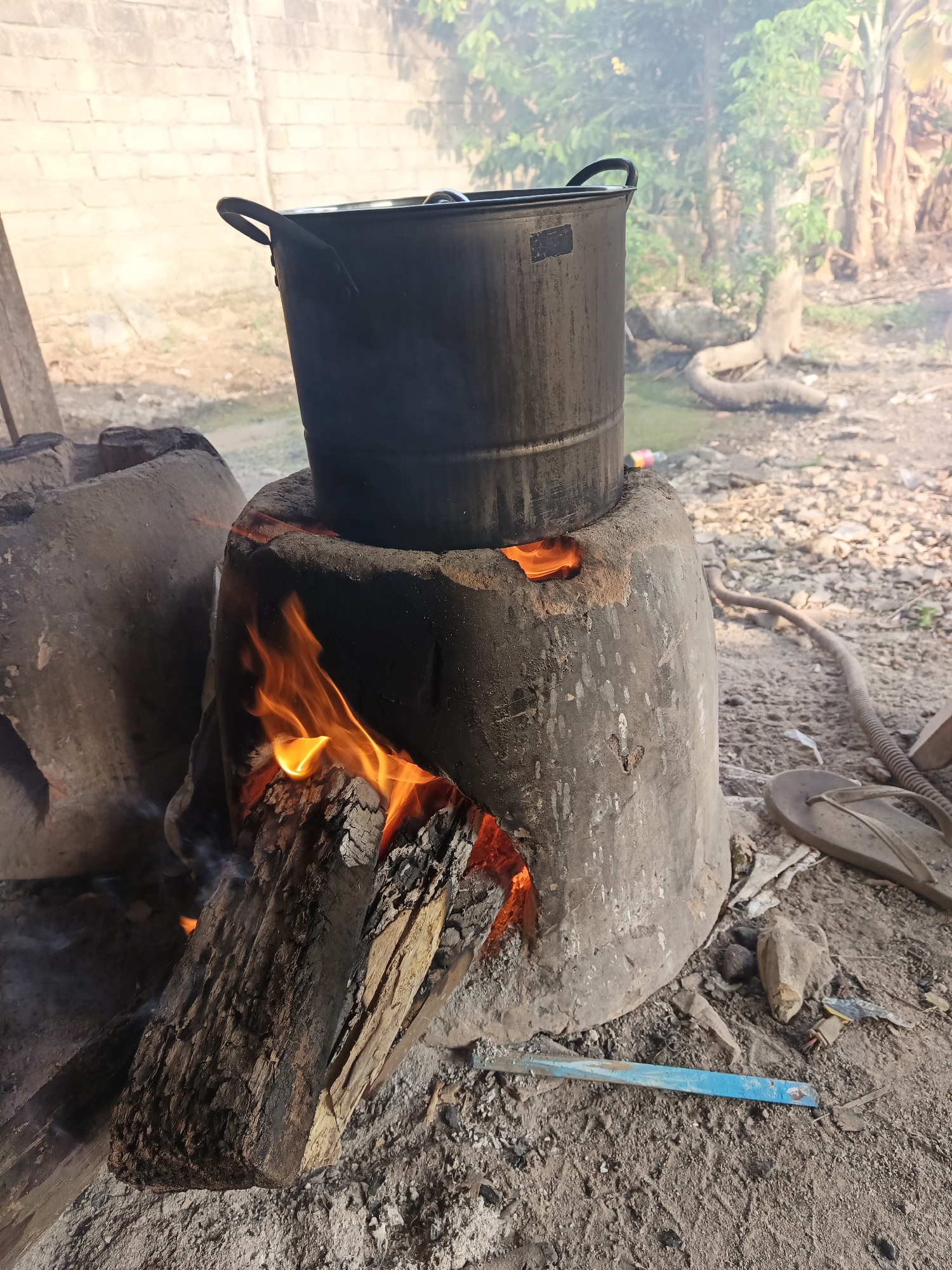 The image depicts an outdoor cooking scene, likely in a rural area of Terbanggi Marga, Indonesia. A large, dark metal pot sits atop a makeshift stove constructed from clay or concrete. Visible flames from burning wood emanate from an opening in the stove, indicating active cooking. Smoke rises from the pot and the fire. The ground appears to be dirt and ash. In the background, there is a brick wall and vegetation, including trees and banana plants, suggesting a tropical or subtropical environment. Scattered debris, including a flip-flop sandal and various pieces of scrap, are visible on the ground. The overall impression is one of a simple, traditional cooking setup in a somewhat unkempt outdoor space.