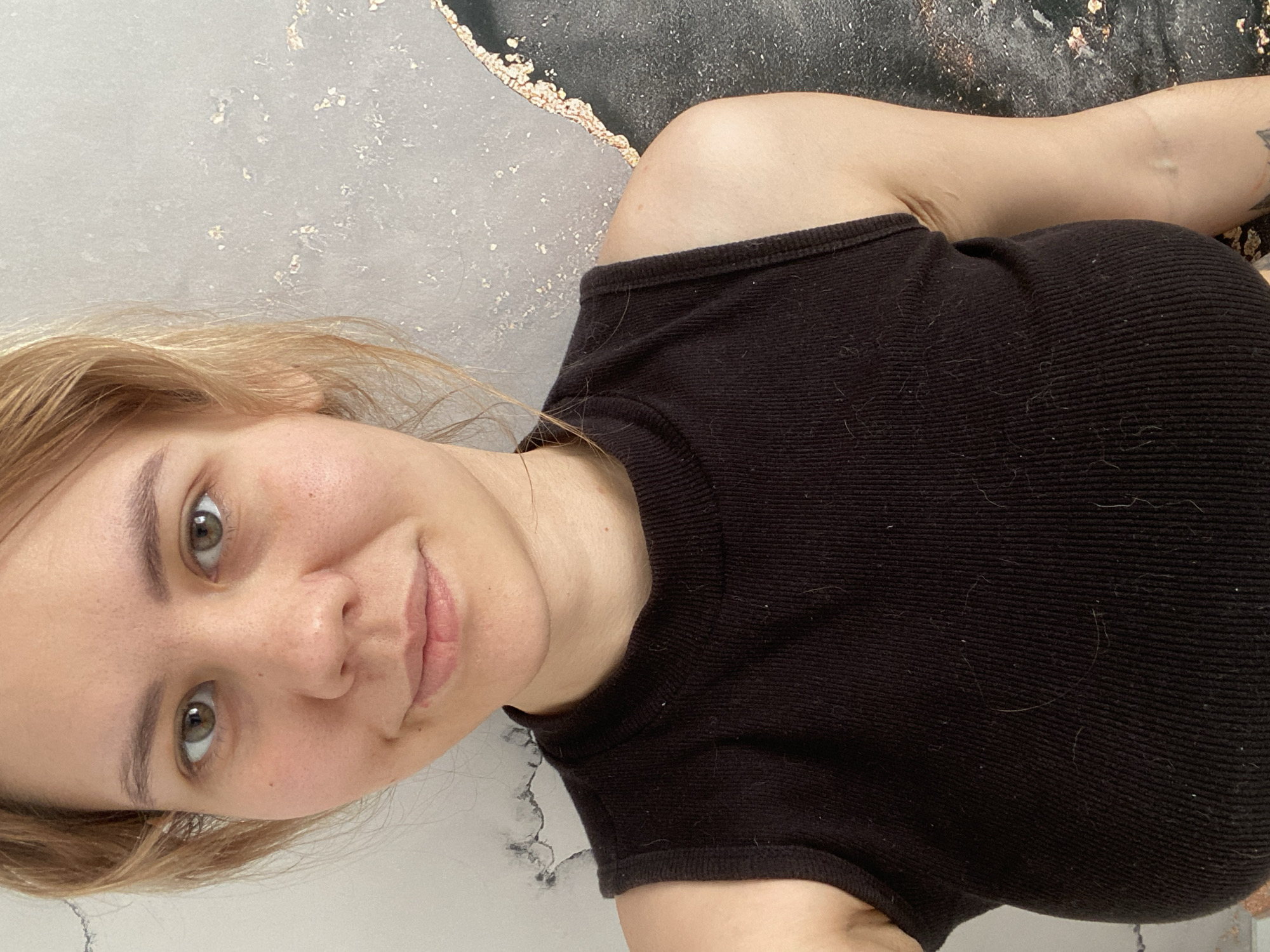 A selfie shows a woman with blonde hair and hazel eyes. She is wearing a black tank top. The background is a mottled gray and black wall, with gold flecks. The photo does not contain location information or objects related to the city of Dyadkovo, Russia.