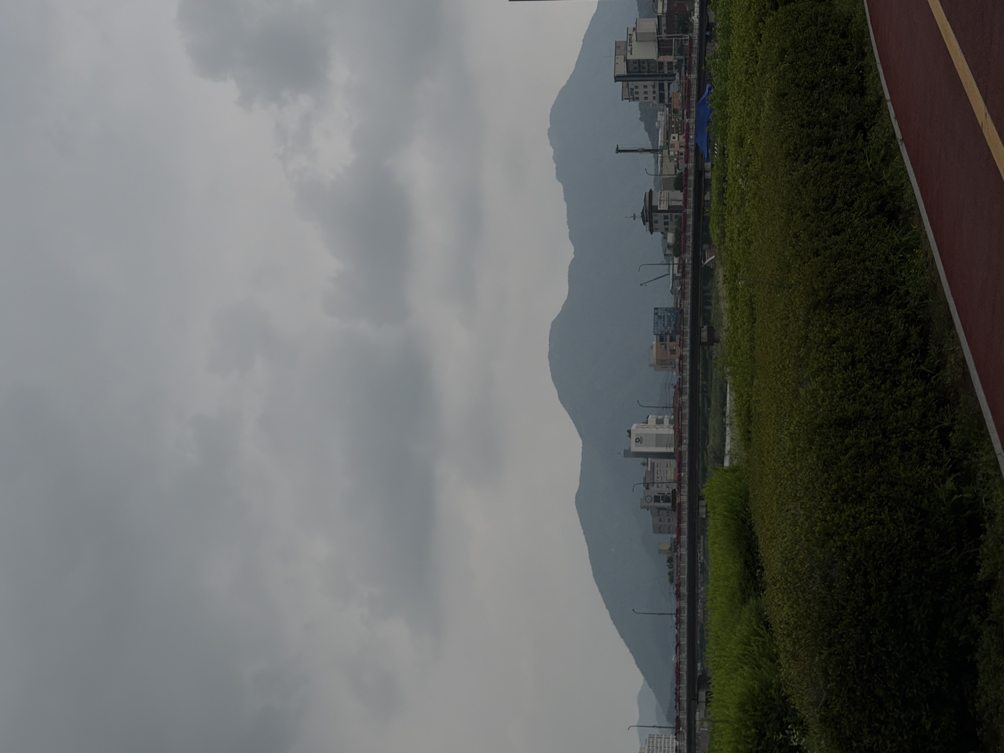 The image depicts a cloudy day in Dongducheon-si, South Korea. In the foreground, a red and yellow striped pathway curves into the frame, bordered by dense green bushes and trees. Beyond the foliage, a wide river flows, with a bridge crossing it in the middle ground. On the far side of the river, a city skyline rises, featuring several multi-story buildings and a distinctive tower-like structure. In the background, a range of hazy mountains dominates the horizon, their peaks softened by the overcast sky. No people are visible in the image.