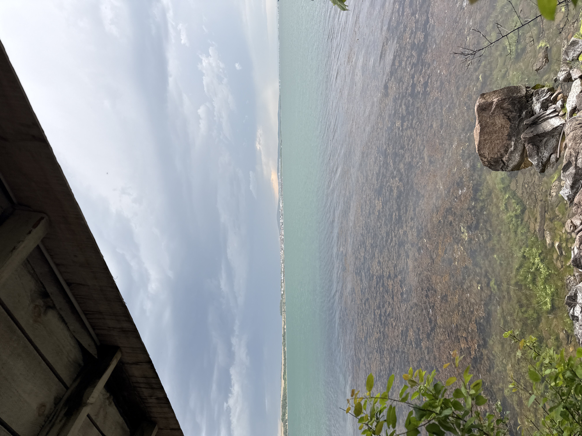 The image presents a view of the Burgas Lake in Burgas, Bulgaria. The scene is divided vertically, with the left side showcasing a cloudy sky and a glimpse of a wooden structure, likely a building or shelter. The right side features the lake's surface, its shallow waters revealing underwater vegetation and a rocky shoreline covered with greenery. In the distance, a faint outline of a cityscape or land mass can be seen across the lake.