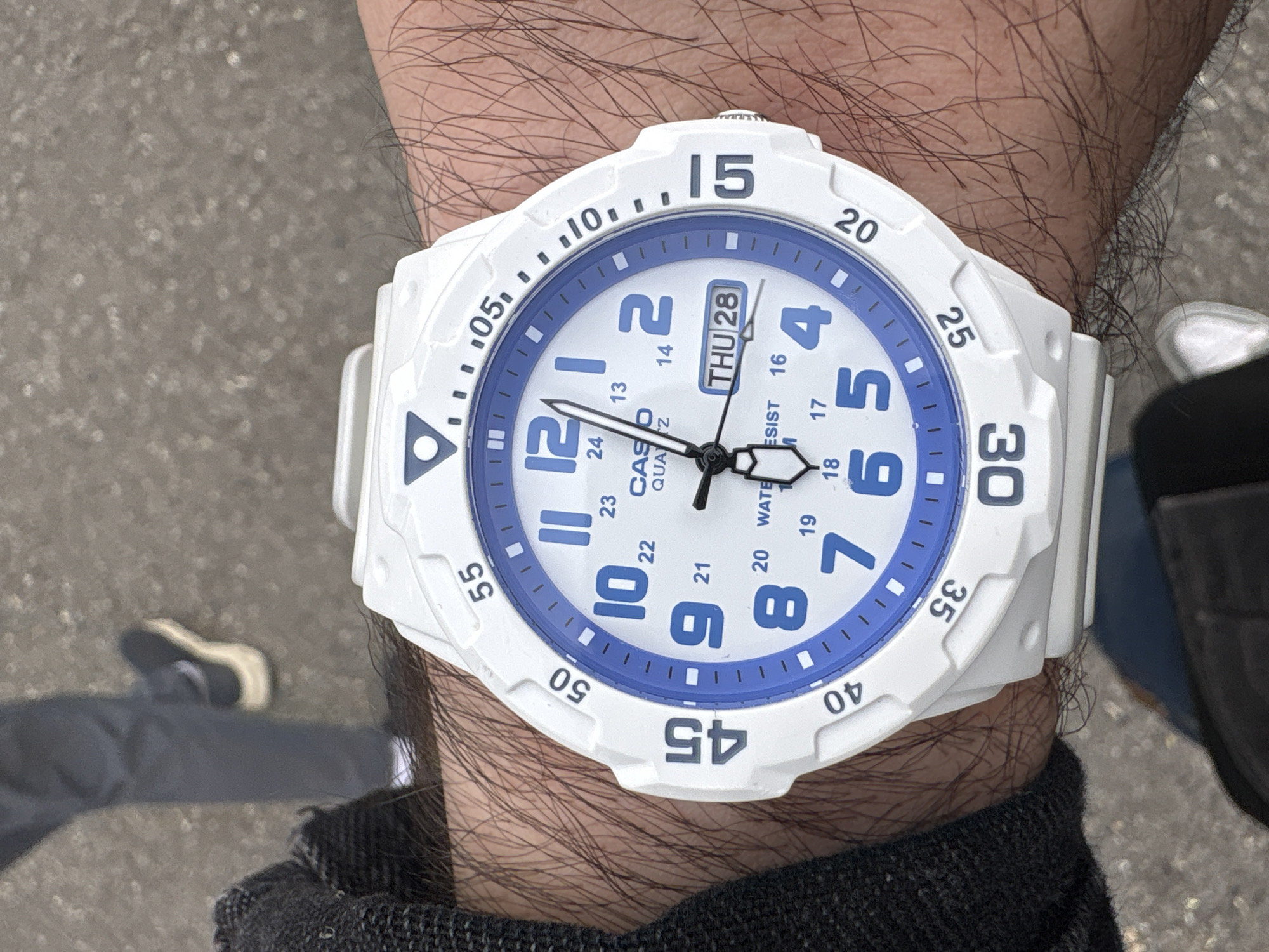 The image shows a person's wrist with a white Casio watch. The watch has a blue and white face with large numbers and a date display showing "THU 28". The wrist is hairy and the person is wearing a black garment. The background is blurred and shows a gray paved surface and a glimpse of clothing and footwear. The location in the city of Yerevan, Armenia is not determinable from the provided image.