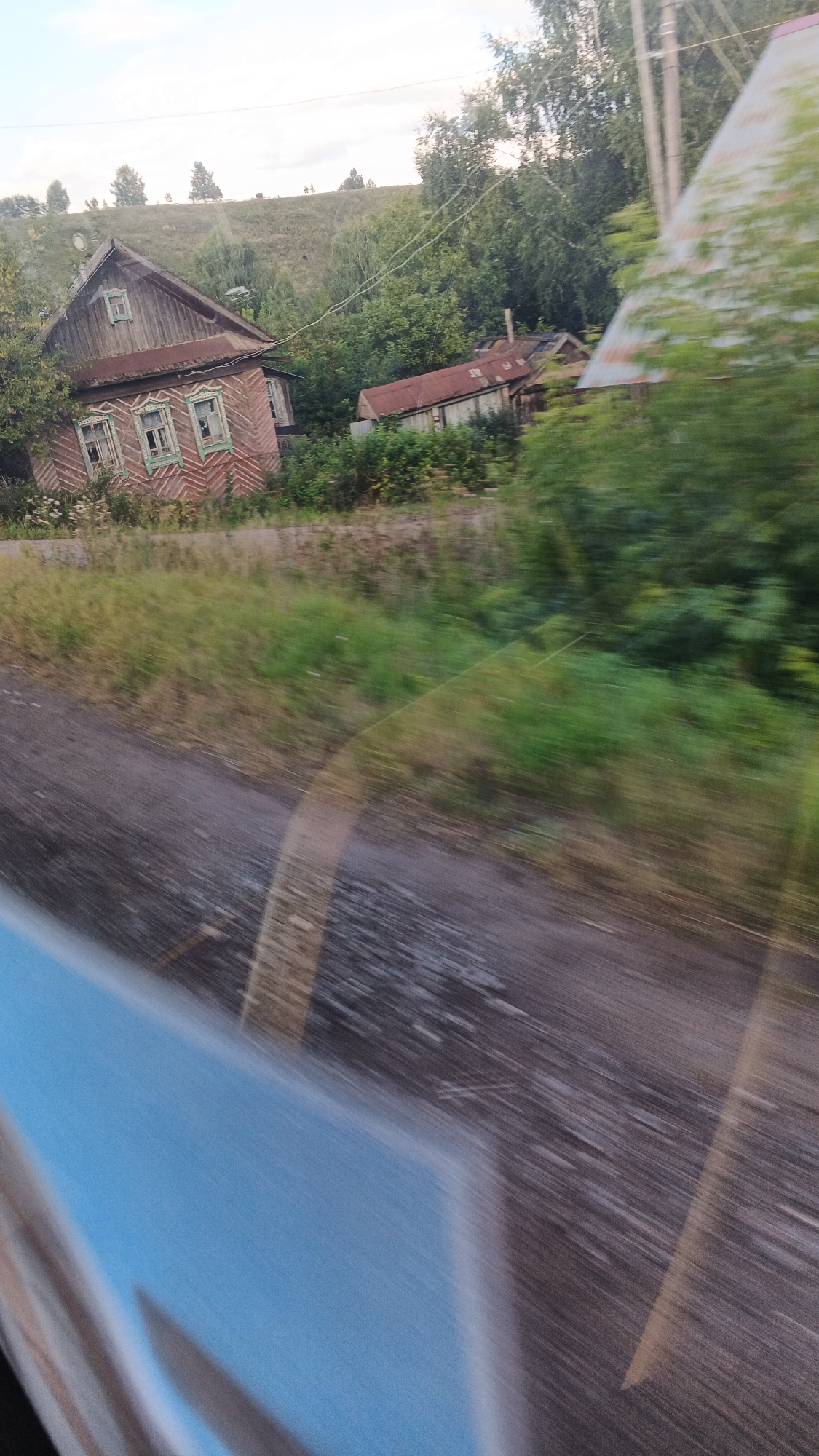 The image shows a rural scene in Buranovo, Russia. Two dilapidated wooden houses with corrugated metal roofs are visible amidst dense green foliage and trees. The houses appear aged and weathered, with peeling paint and broken windows. A dirt road runs in front of the houses, and the foreground is blurred due to motion, suggesting the image was taken from a moving vehicle. A blue object, possibly a part of the vehicle, is partially visible in the bottom left corner. The sky is overcast.