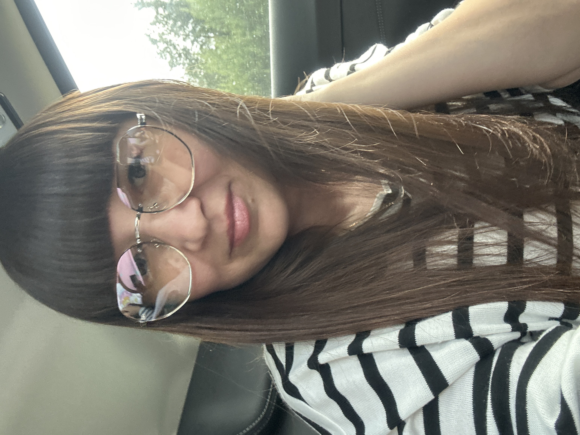 The image is a selfie of a woman with long brown hair and bangs, wearing glasses and a black and white striped shirt. She is sitting in what appears to be the passenger seat of a car. The background shows trees and a bright sky, suggesting an outdoor setting, possibly in a rural or suburban area. There are reflections in her glasses, showing parts of the car's interior and possibly other people. The woman is looking towards the camera and her right hand is resting on her neck. The lighting suggests it is daytime. The location of Dokshitsy, Russia is not depicted in the image.