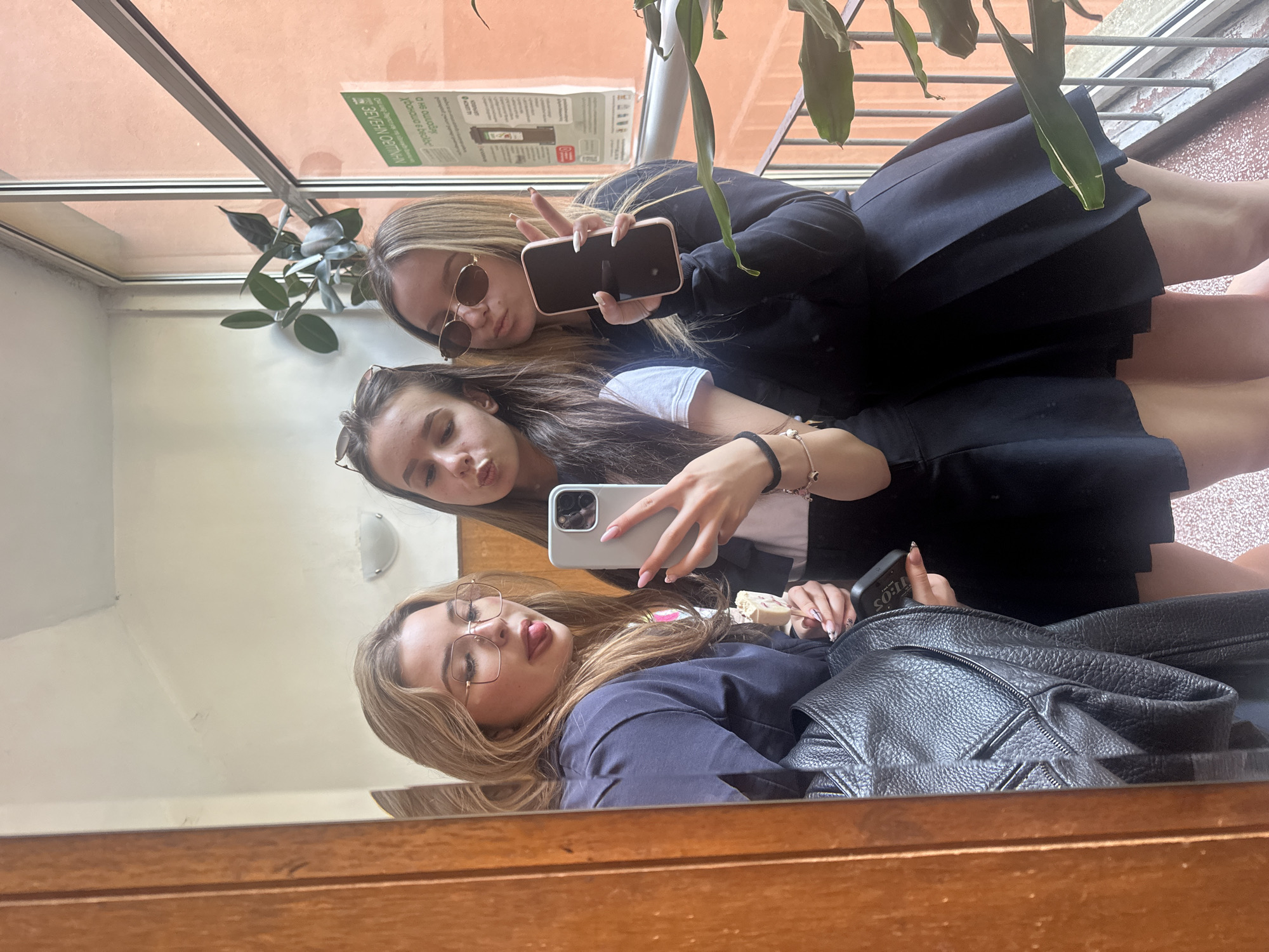Three young women are taking a selfie in a mirror. All are wearing skirts, likely school uniforms. The woman at the top is wearing sunglasses and holding her phone, making a peace sign. The woman in the middle is holding a phone as well. The woman at the bottom is wearing glasses and has her tongue sticking out. A black leather backpack is visible near the bottom of the frame. In the background, there is a window with a plant and a poster in Bulgarian, identifying the location as likely being in Blagoevgrad, Bulgaria.