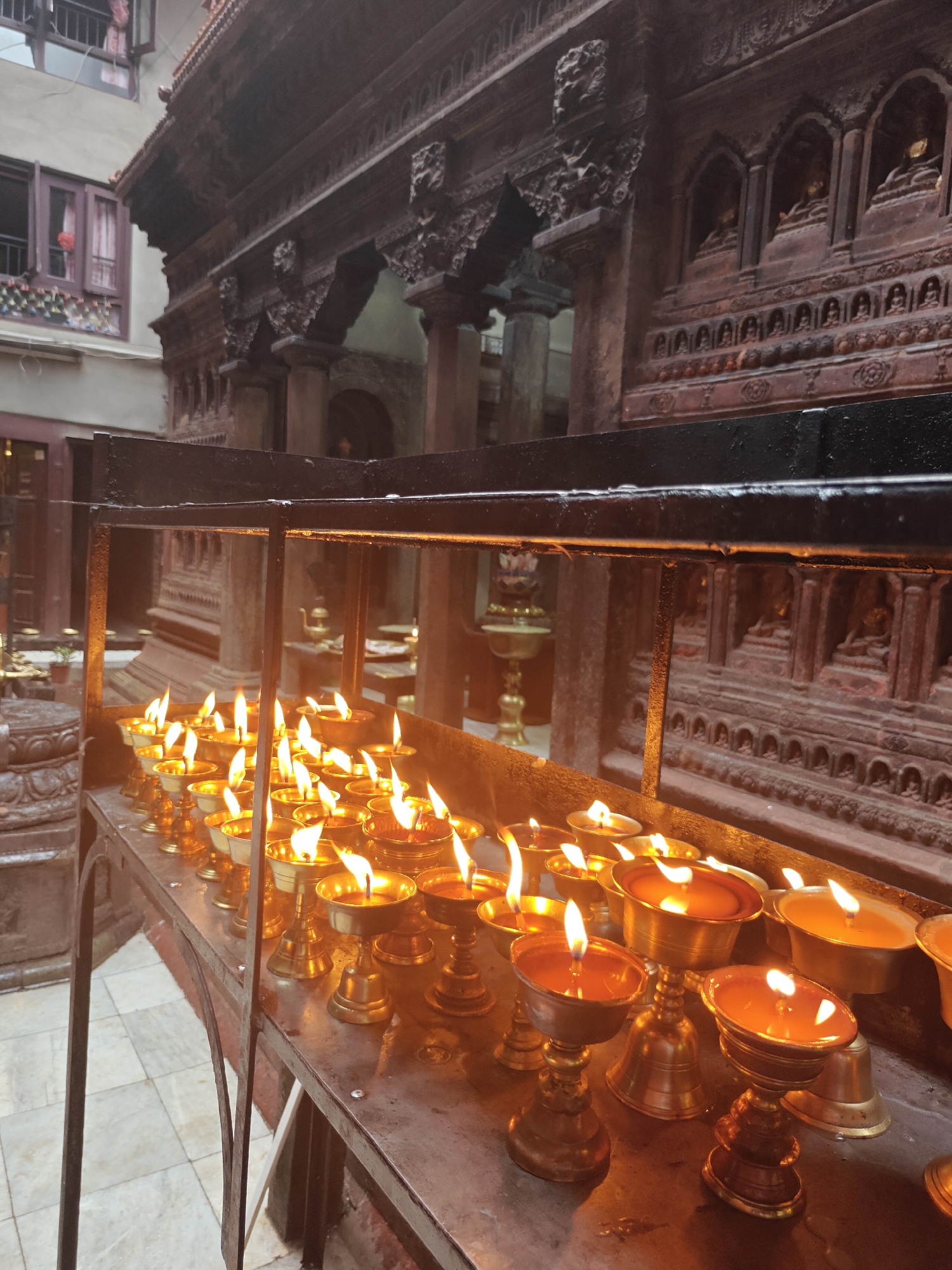 The image depicts a close-up view of numerous brass oil lamps, each with a lit wick, arranged on a tiered metal shelf. The lamps are of varying heights and designs, with some featuring decorative bases and others being simpler in form. The flames cast a warm, orange glow on the surrounding surfaces, reflecting off the polished brass. In the background, a richly carved wooden structure, possibly part of a temple or shrine, is visible. This structure has intricate carvings and rows of small niches that appear to contain figurines. To the left of the wooden structure, a portion of a building with windows is visible. The floor in the foreground is tiled. The overall scene suggests a spiritual or devotional setting, likely within a temple complex in Lalitpur, Nepal, given the architectural style and the presence of prayer lamps. No people are visible in the frame.