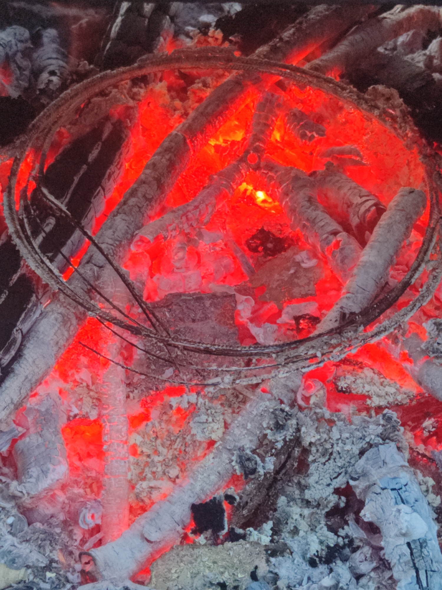 This is a close-up, overhead shot of a dying campfire. The main subjects are the embers and ashes of burnt wood, with several logs partially visible. There is also a circular metal object, possibly part of a grill or fire pit, positioned towards the top of the frame, encircling some of the hottest coals. The scene is dominated by the glowing red of the embers, which intensely illuminate the surrounding ash and wood. The wood appears charred and broken, indicating it has been burning for some time. The ash is thick and powdery, with some larger, chunkier pieces. The metal ring shows signs of heat and wear. The lighting is entirely from the fire itself, creating a dramatic contrast between the bright reds and the dark shadows. There are no people, text, or other elements to suggest a specific time of day, weather, or precise location beyond a general outdoor setting where a fire would be. The overall impression is one of intense heat and the remnants of a fire.