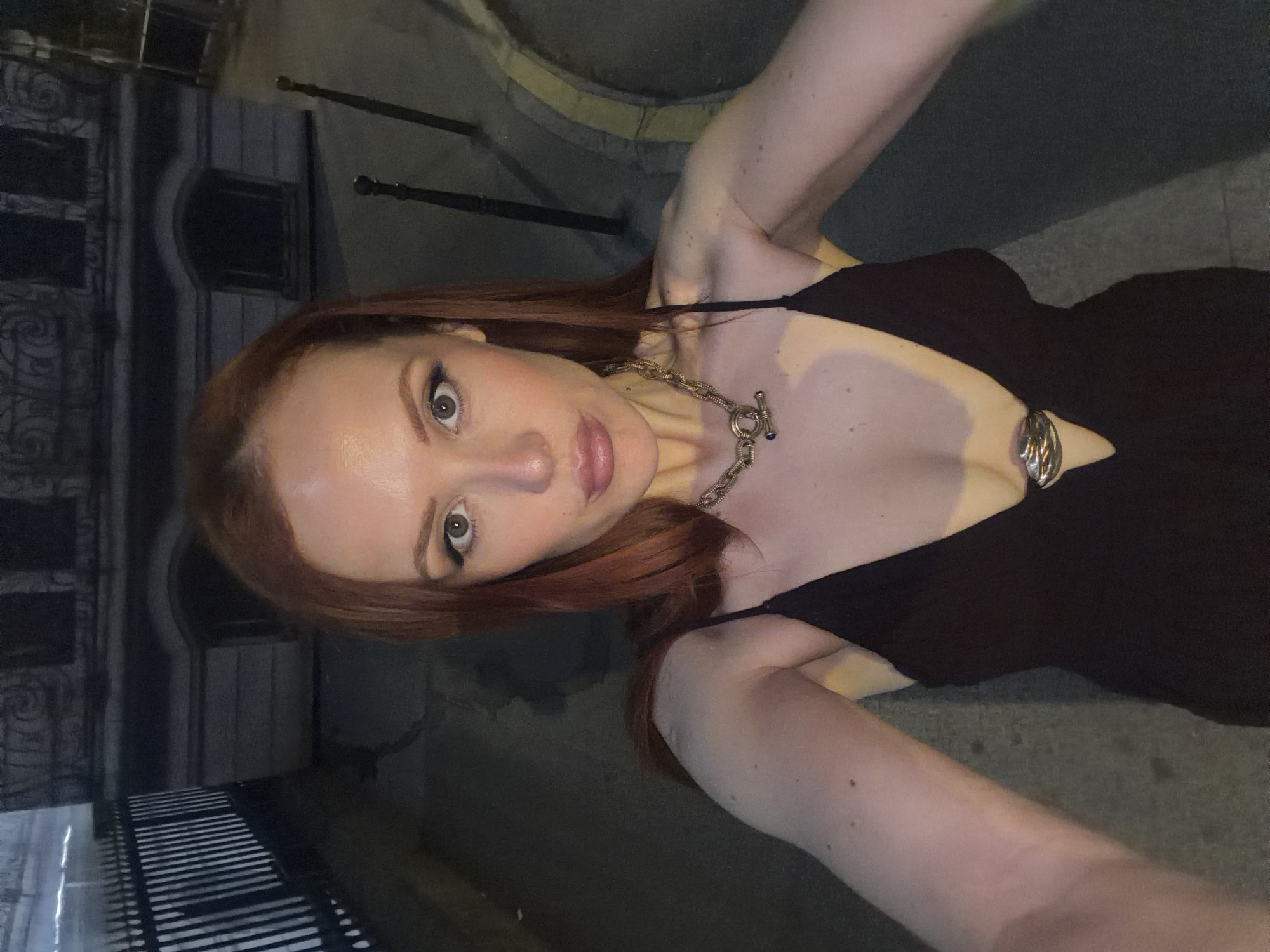 A woman with long, auburn hair poses for a selfie at night in Paris, France. She is wearing a black dress with a V-neck and a chunky silver chain necklace. The background includes wrought iron fencing, a building facade with arched windows, and a street with a curb. The woman's skin tone is fair. The lighting suggests a mix of natural and artificial sources.