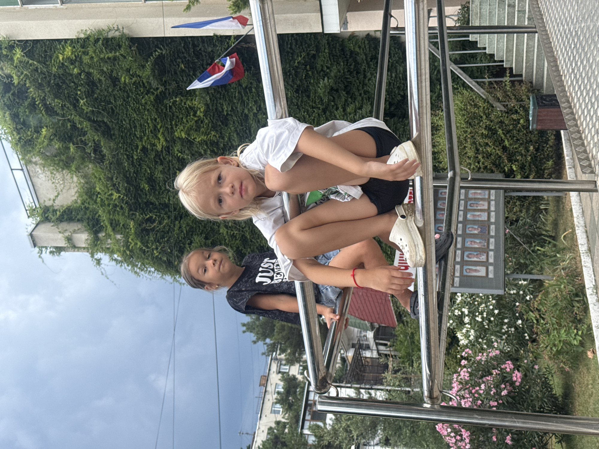 Two young girls, one with blonde hair and wearing a white t-shirt and black shorts, and another with brown hair wearing a black t-shirt and denim shorts, are sitting on a metal railing. They are outside, with a building covered in green ivy in the background. Two flags, one with red, white, and blue stripes and another with blue and white stripes, are visible on the building. There are pink and white flowers in the foreground. A set of stairs and a ramp are also visible. The sky is overcast. The location is identified as Partenit, Ukraine.