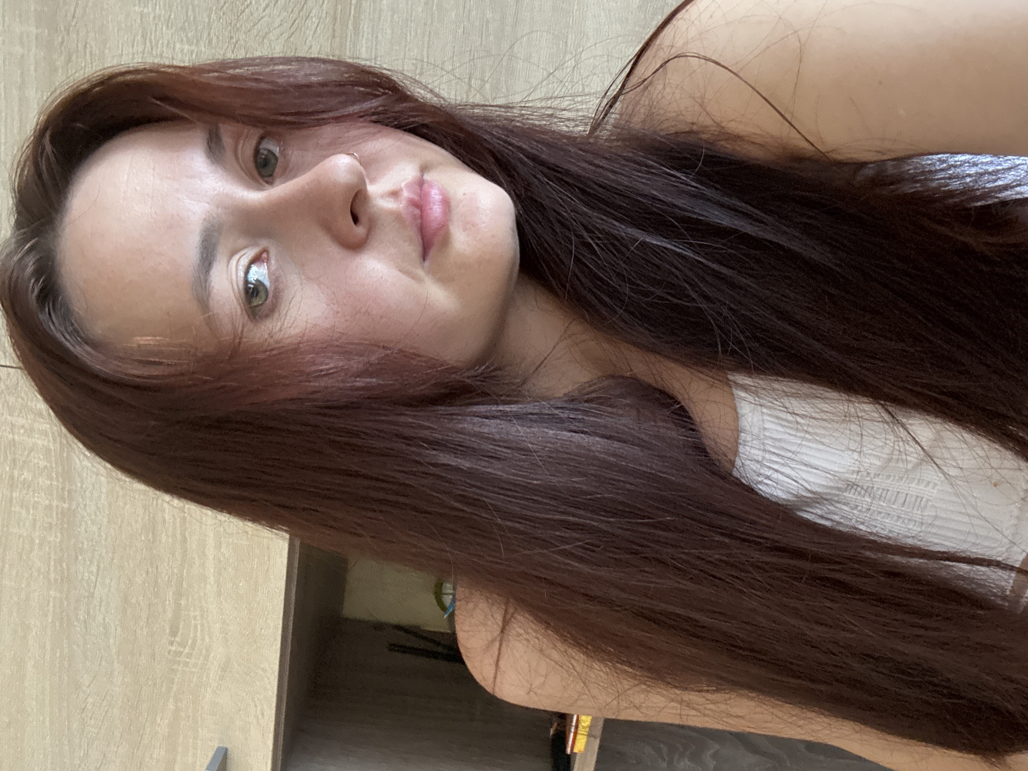 A young woman with long, dark brown hair and green eyes is pictured from the chest up. She is wearing a white ribbed tank top. She has a small nose ring. The background appears to be a light-colored wooden surface. The image is taken from a slightly high angle. No specific location in Naberezhnye Chelny, Russia is identifiable.