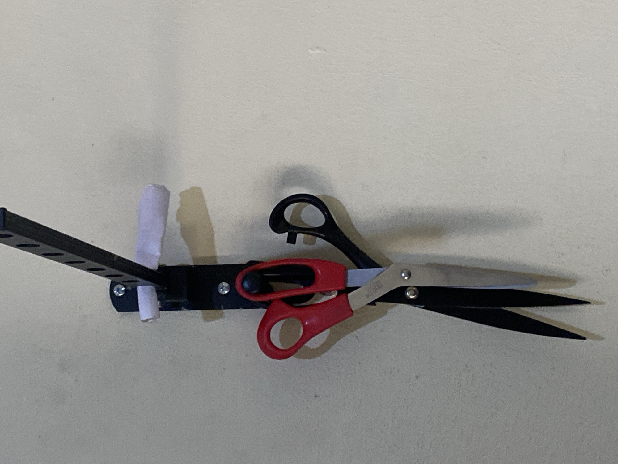 A pair of red-handled scissors and a pair of black-handled scissors are hanging from a black metal rack mounted on a textured, light-colored wall. The rack is attached to the wall with screws. A small roll of white material, possibly tape, is also attached to the rack. The image is an indoor shot, likely in a home or workshop. There are no people present in the image. The location is not specified as Jauro Yinu, Nigeria.