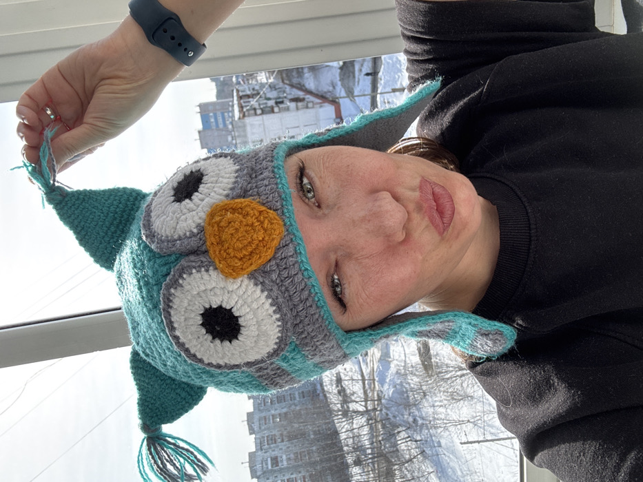 A front-facing photograph captures an adult female from the chest up, positioned indoors looking out through a window onto a snow-covered urban environment in Chaikovsky, Russia. The female has light-colored eyes and visible freckles. She is wearing a black long-sleeved garment and a crocheted owl-themed winter hat predominantly teal, grey, white, and orange, which features large white and black eyes, an orange beak, and ear flaps. Her left hand is raised, grasping a teal and grey tassel extending from the top right side of the hat. A dark-colored smartwatch is visible on her left wrist. The subject's lips are puckered. In the background, outside the window, multiple multi-story residential buildings and bare trees are visible, with ground covered in snow under a bright sky, indicative of a winter or early spring setting. White window frames are present along the top and right edges of the image.