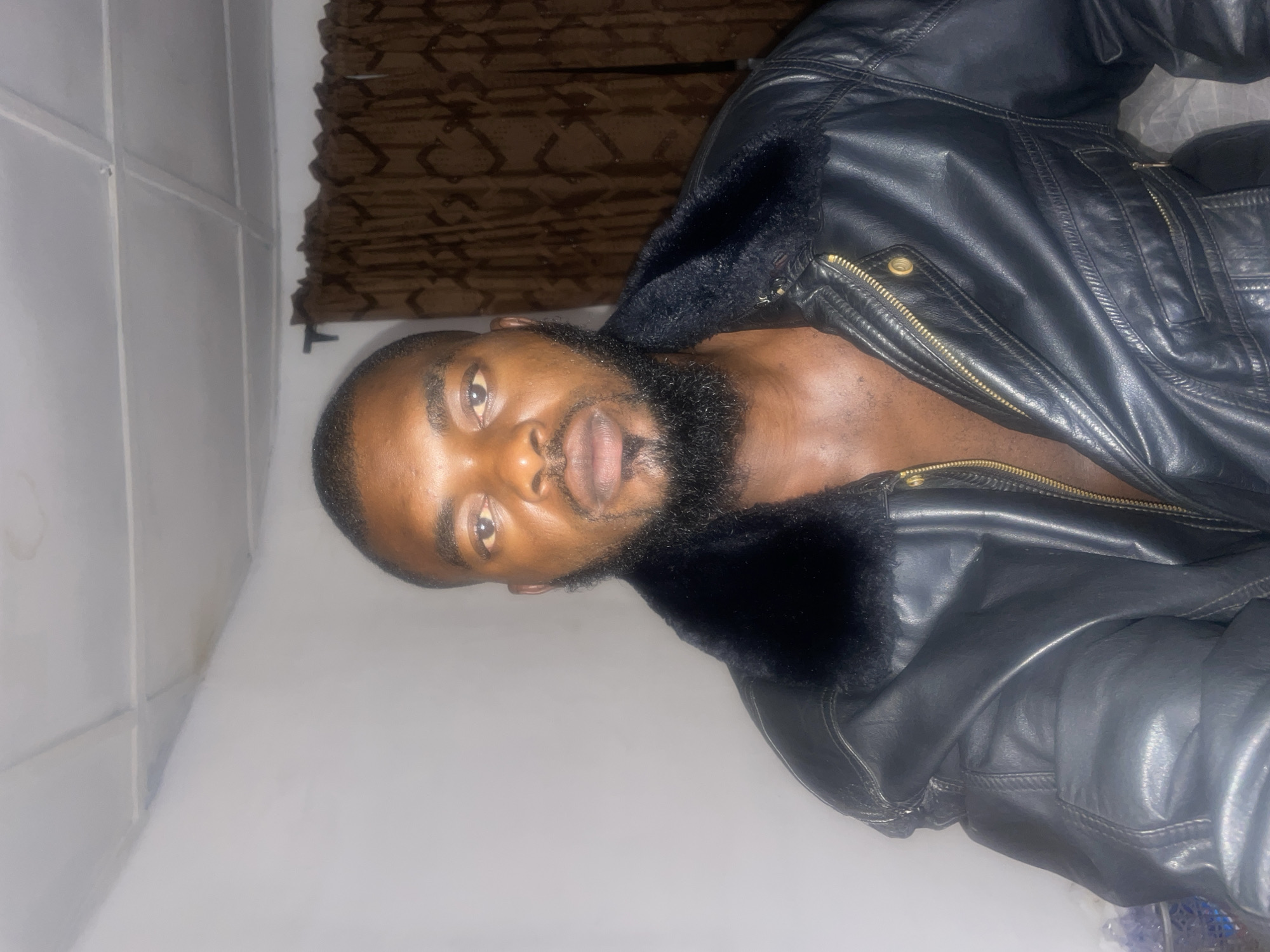 A person with dark hair and a full beard is wearing an open black leather jacket with a dark fur collar. The person is facing forward and looking at the camera. The background consists of a textured brown window treatment and a light gray wall. There is no discernible information to determine if the location is in Jos, Nigeria.