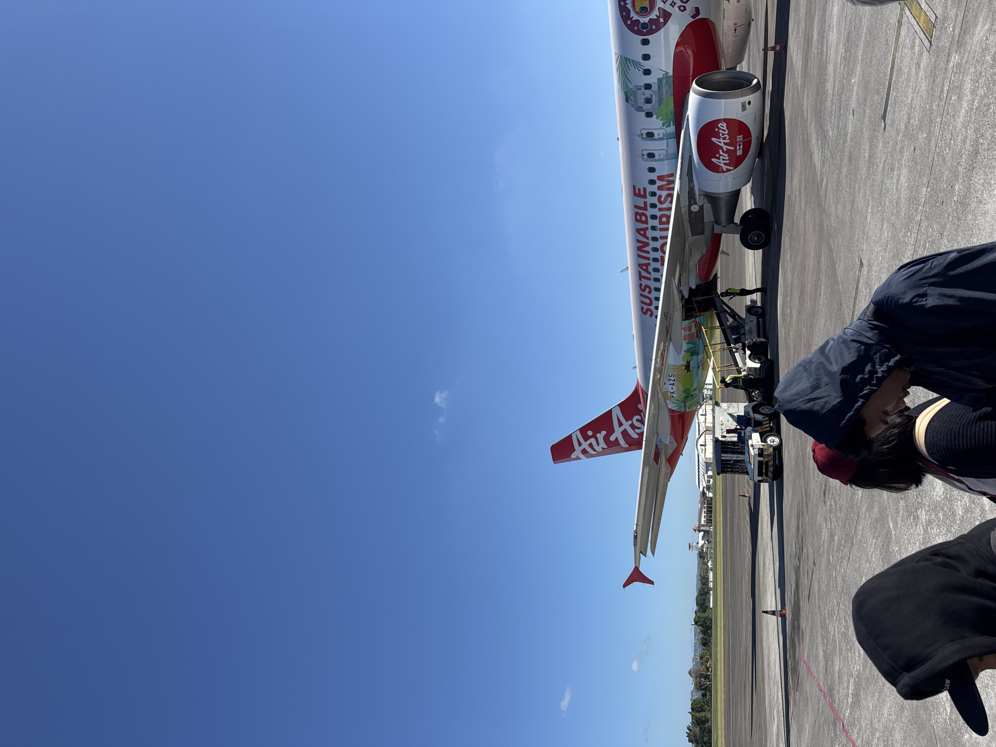 The image shows an AirAsia airplane on an airport tarmac under a clear blue sky. The plane is predominantly red and white and features the text "SUSTAINABLE TOURISM" printed along its fuselage, along with "Air Asia" on the engine. The airplane's tail is visible, also displaying the "Air Asia" logo. Several people are in the foreground, their backs to the camera, suggesting they are observing or waiting near the aircraft. One person wears a dark hooded jacket and a red cap. Another person in the foreground wears a black hooded jacket and a dark baseball cap. A baggage tug vehicle is parked near the plane, and service equipment is also present. The scene appears to be in a warm, sunny location, possibly during the day, given the bright lighting and clear sky. The presence of trees and a distant treeline in the background indicates an outdoor setting, likely an airport in Kuta, Indonesia.