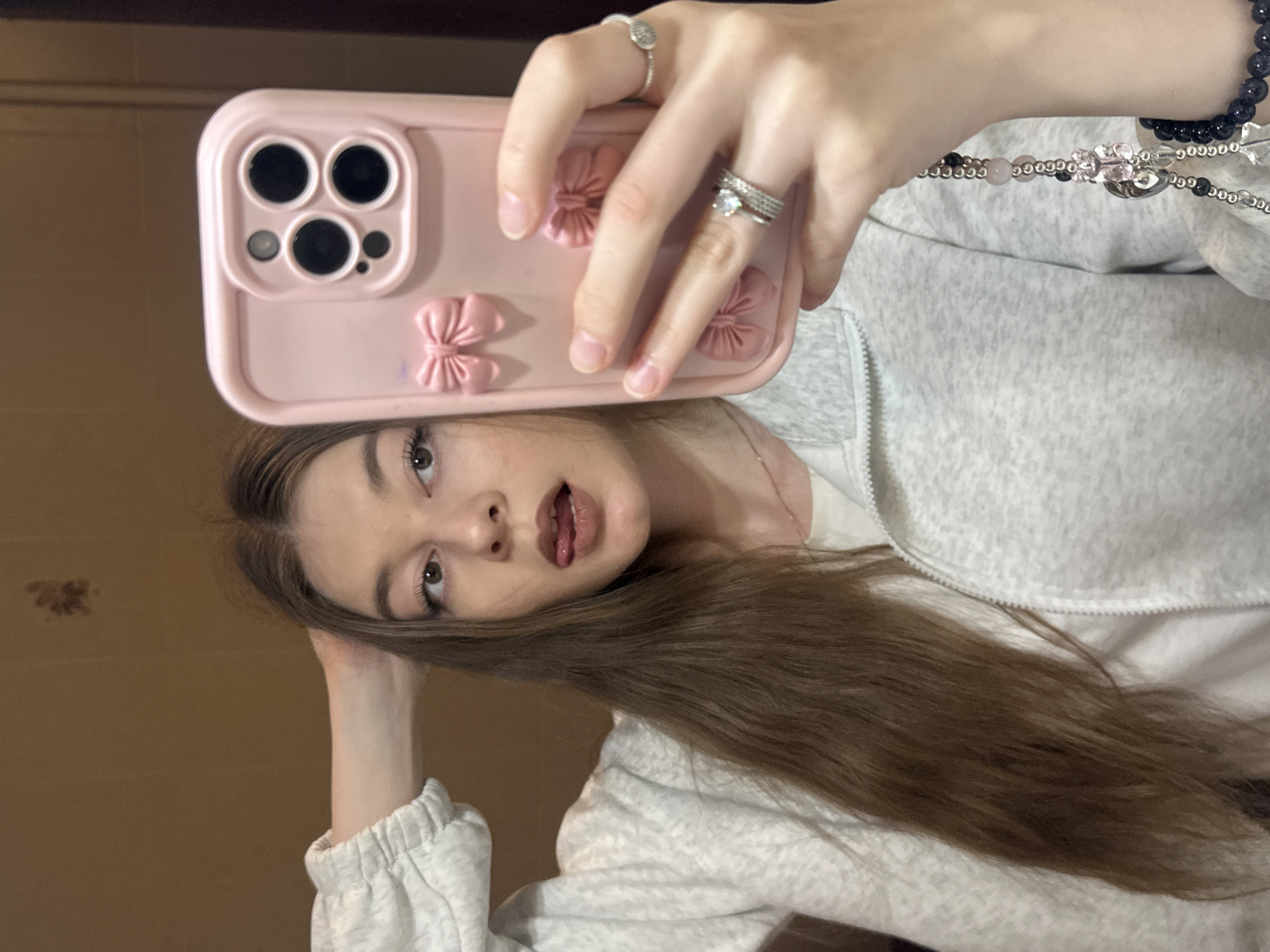 A young woman takes a selfie using a pink phone case adorned with bow details. She wears multiple rings on her hand holding the phone. She has long brown hair and is wearing a light gray zip-up jacket over a white shirt. A beaded necklace is visible around her neck. The background appears to be a neutral-toned wall. There is no indication of the location being Kazan, Russia.