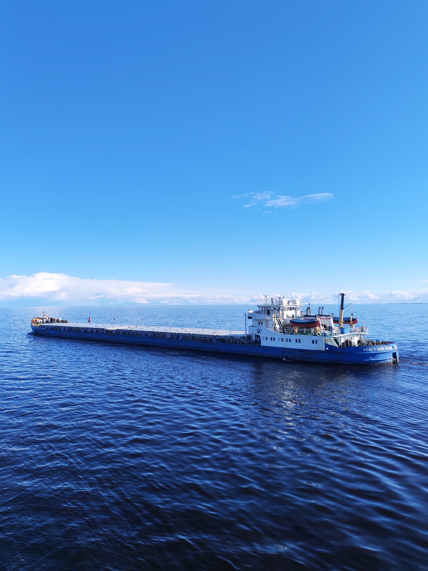 The image depicts a long, blue and white cargo barge sailing on a deep blue body of water under a bright blue sky with scattered white clouds. The barge has a multi-level white superstructure at its stern. The name "ВОЛГО-ДОН 5060" is painted on the stern of the ship in white letters. The image does not contain any visual information that would allow the determination of the specific location in Novaya Svobodka, Russia. The focus is solely on the barge on the water. There are no visible people or actions occurring.
