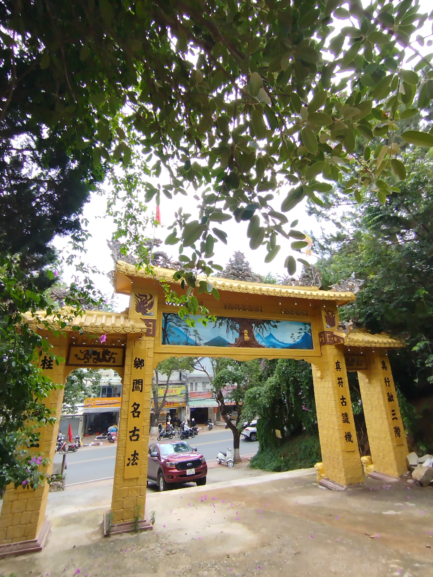 Here is a detailed description of the image:
**Scene Overview:** The image captures a view of a golden, ornate gate. The gate is positioned on a slightly elevated platform. The other side of the gate opens onto a street scene in Lâm Viên - Đà Lạt, Vietnam.
**Gate Details:** The gate is constructed from what appears to be textured, yellow stone or concrete. The structure is decorated with Chinese characters and artwork, particularly a scenic mountainscape.
**Street Scene:** A red Ford vehicle is positioned within the gate's view. Further down the road, multiple motorcycles and other vehicles are visible. The street is lined with buildings, including several businesses with signage visible in Vietnamese.
**Environment:** The scene is framed by trees and lush vegetation, creating a sense of nature integrating with the urban landscape. The sky appears overcast, casting soft lighting across the scene.
**Additional Details:** Purple flower petals are scattered on the pavement in the foreground. The condition of the gate suggests it's a well-maintained landmark. The overall composition balances architectural details with the everyday life of the surrounding city.