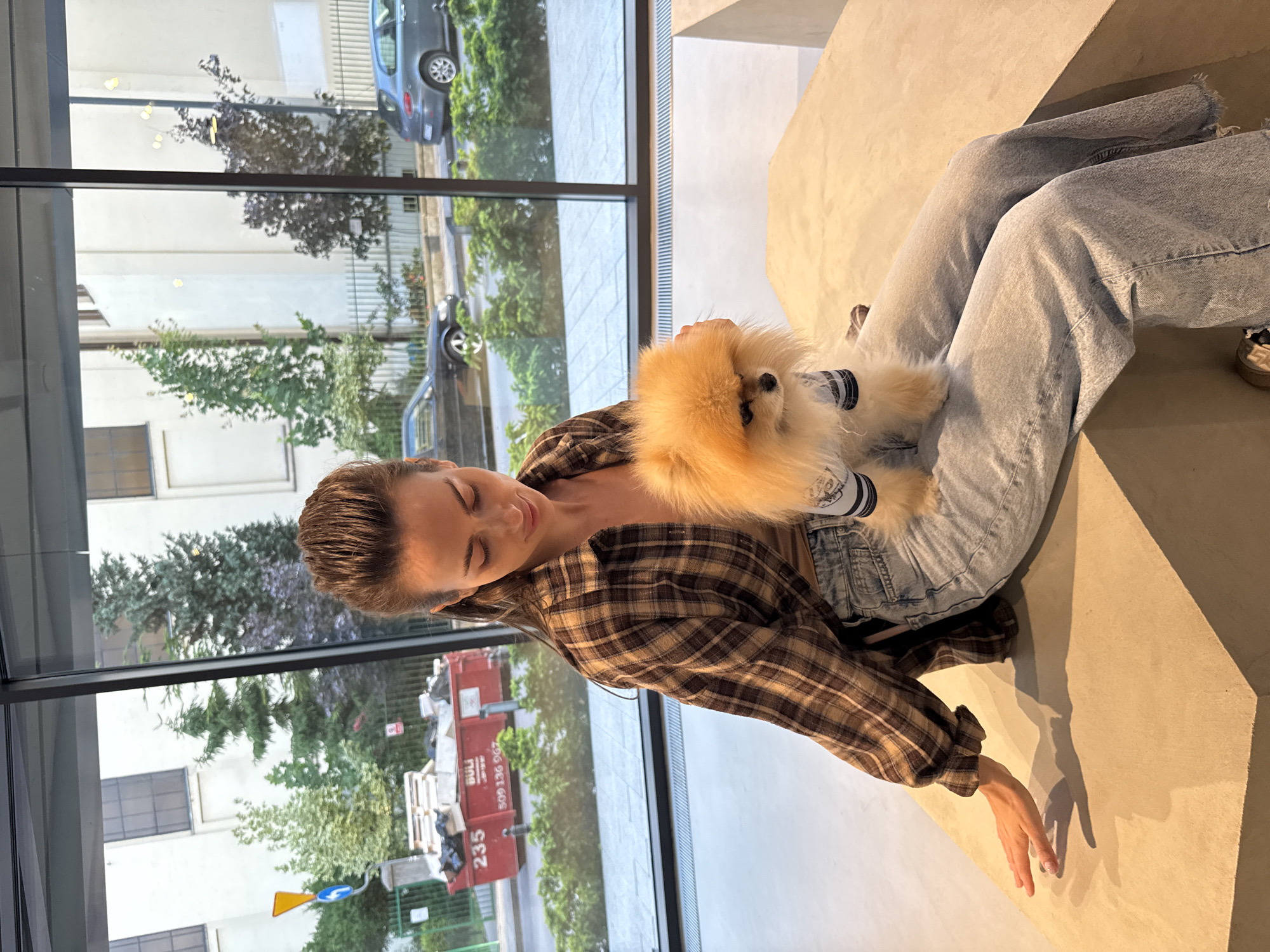 A woman sits in a relaxed pose indoors, holding a small fluffy dog on her chest/lap, beside a large window overlooking an outdoor urban scene. The woman has dark hair pulled back and is wearing a brown and black plaid long-sleeved shirt over a lighter top, paired with light-wash, ripped jeans. Her eyes appear closed or are cast downwards, suggesting a moment of quiet contentment.
The dog, a light brown/orange Pomeranian, is wearing a white shirt with black horizontal stripes and is nestled comfortably against the woman. Its fur is very fluffy, and it appears calm.
The indoor setting features a light-colored, possibly concrete or stone, surface that the woman is sitting on, which extends into the background. Ample natural light streams in through the large windows.
Beyond the windows, an outdoor environment is visible, featuring various buildings with white and light-colored facades, green trees, and parked cars, including a dark-colored sedan. In the foreground of the outdoor scene, a large red skip or dumpster is prominent, with white text reading "BULI," "509 136 907," and "235." A yellow and blue road sign is also visible near the bottom left of the outdoor view. The overall lighting suggests it is daytime, possibly sunny, creating shadows on the indoor surface.