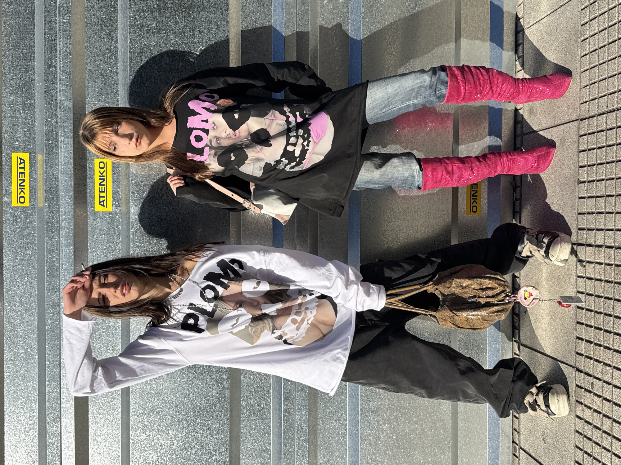 Two young women are lying on steps made of textured gray metal. Both are wearing long-sleeved shirts with graphic designs and text. The woman on the top is wearing a black shirt with a pink design and light-wash jeans, paired with tall, glittery pink boots. The woman on the bottom is wearing a white shirt with a graphic design and black pants, along with sneakers. She has a brown snakeskin-patterned purse. Both women are facing the camera. "ATENKO" is printed on yellow tape on the steps. The background includes a section of concrete sidewalk with small, square tiles. The location is Buenos Aires, Argentina.