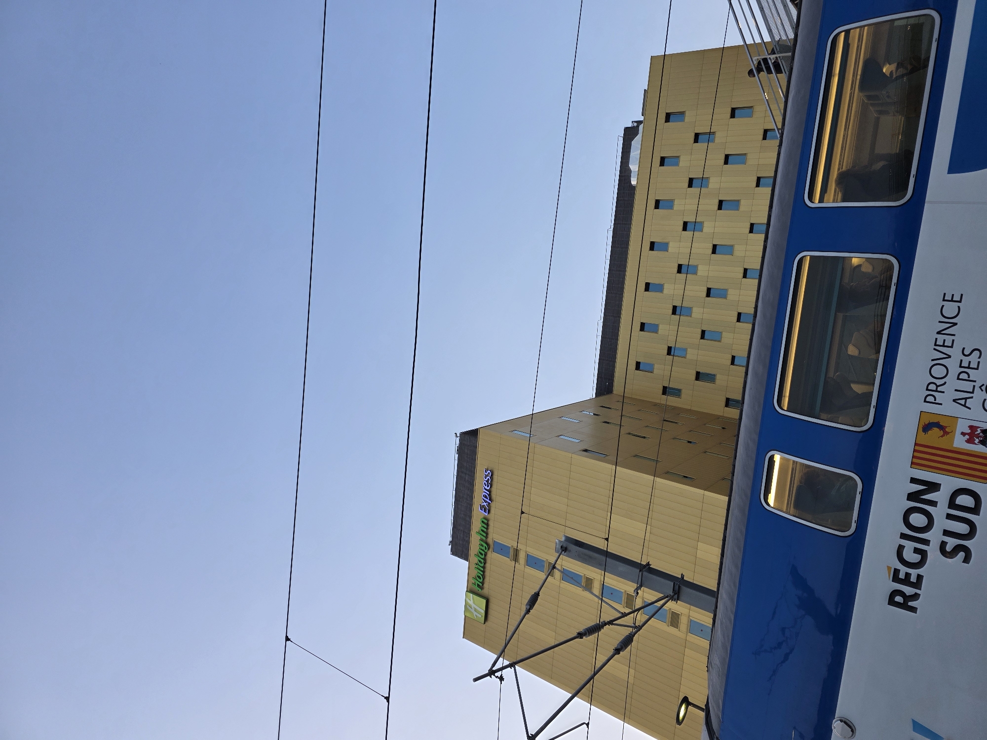 The image captures an urban scene in Nice, France, with a focus on a modern, golden-colored building bearing the "Holiday Inn Express" logo. In the foreground, a blue and white train car is visible, with "REGION SUD" and "PROVENCE ALPES" text and emblems on its side, indicating it's a regional train. The train has illuminated windows, suggesting passengers inside. Overhead, power lines for the train tracks crisscross the clear blue sky. The building's facade is punctuated by numerous small, rectangular windows. A person is visible on a walkway or bridge near the top right of the building. The perspective is looking upwards, emphasizing the height of the building and the overhead infrastructure.