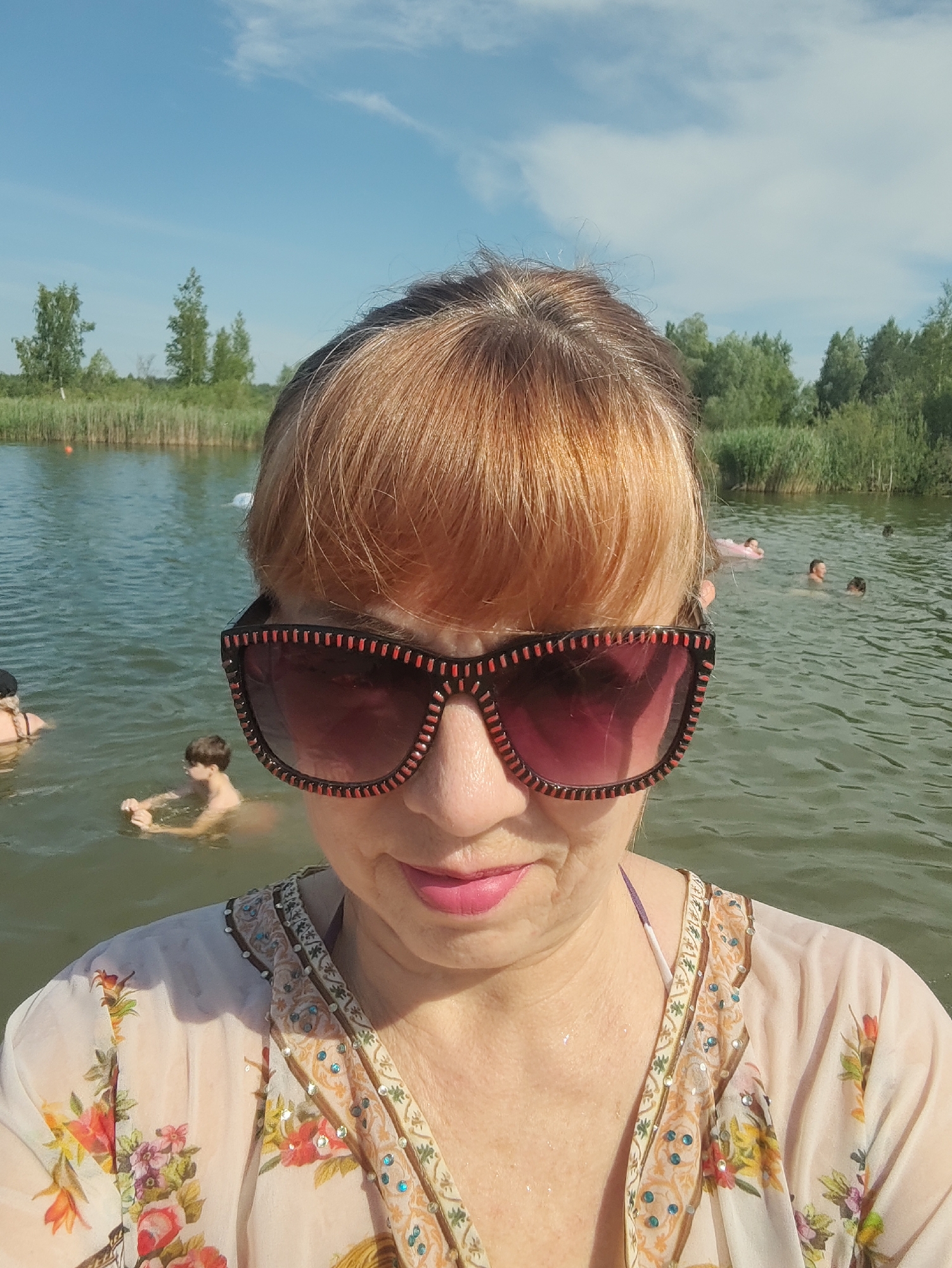 This is a portrait-oriented, eye-level, medium shot photo taken on a sunny day at a lake in Rovnoe, Russia. The subject, a woman, is in the foreground, facing the camera and wearing oversized sunglasses and a flowered blouse over a bikini. Her hair is styled with bangs. In the background, other people are swimming in the lake, with some in the distance on inflatable toys. The lake is surrounded by trees and vegetation under a clear blue sky with scattered clouds. The water appears calm and the overall mood is relaxed and summery. The sunlight suggests it is daytime. No visible text is present in the image.