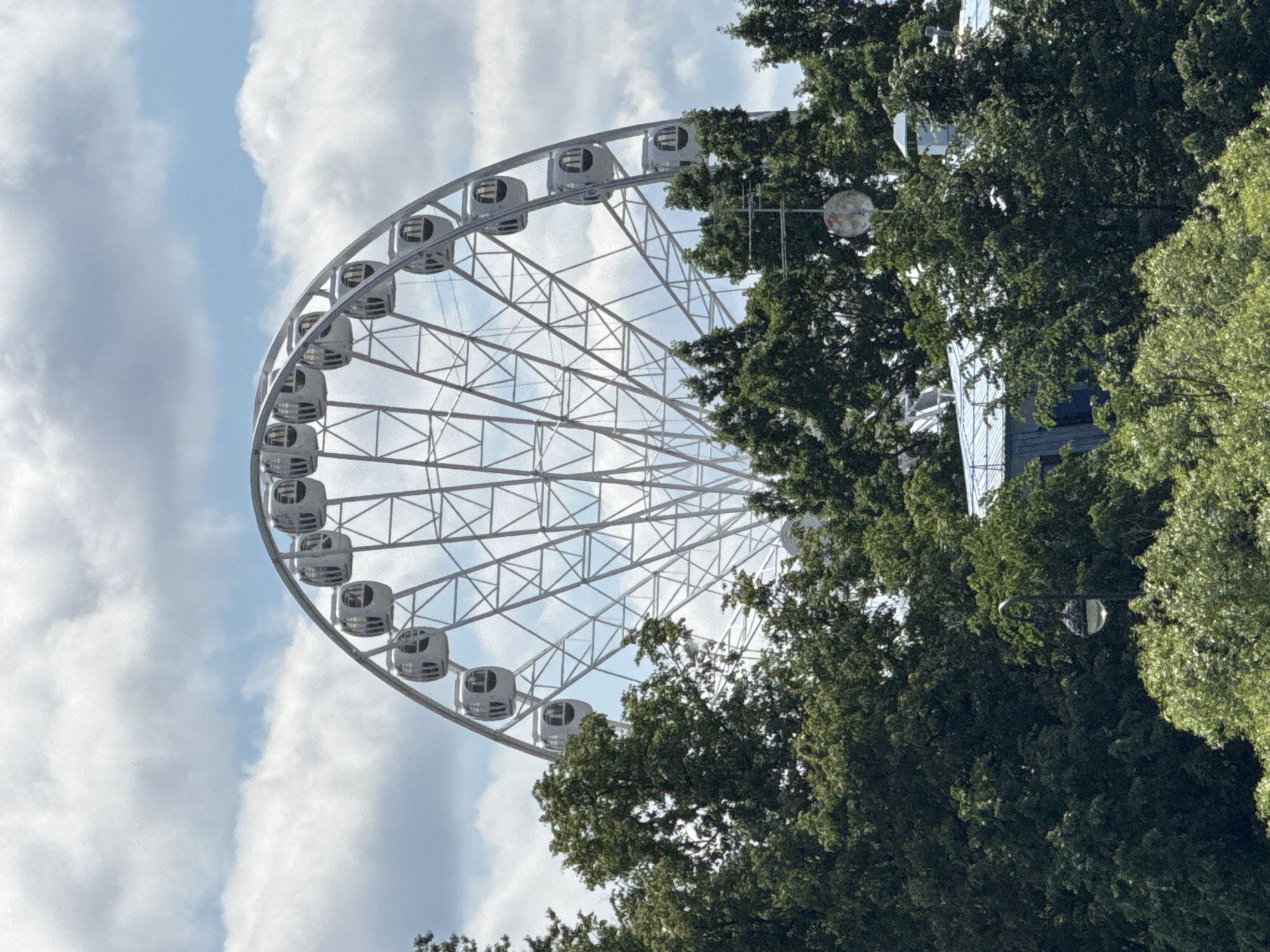 The image shows a Ferris wheel in the city of Saint Petersburg, Russia, with lush green trees in the foreground obscuring part of the wheel and the surrounding environment. The Ferris wheel is white and appears to be made of metal, with numerous cabins. The sky is cloudy with patches of blue visible. A street lamp is visible among the trees. The architecture of some buildings can be seen peeking through the foliage. The overall scene suggests a sunny day in a park or recreational area.