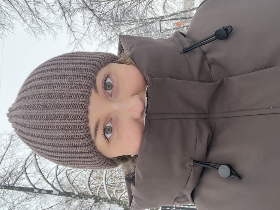 The image displays a close-up, chest-up portrait of an individual, likely female, photographed outdoors in winter conditions. The subject wears a brown ribbed knit beanie and a brown, high-collared winter jacket, with the collar pulled up to obscure the mouth and chin. Visible facial features include greenish-hazel eyes, a nose, and the upper cheeks. The eyes are open and directed forward, appearing to look into the camera. The jacket collar features black bungee cord drawstrings with circular black plastic toggles on both sides. The background reveals a winter landscape of bare tree branches lightly coated in snow or ice, set against an overcast, light gray sky. Indistinct urban elements, such as distant buildings or streetlights, are faintly visible, suggesting a park or urban area. No specific landmarks are present within the image to precisely identify a particular location within the city of Chaikovsky, Russia. The overall environment indicates cold, snowy weather.