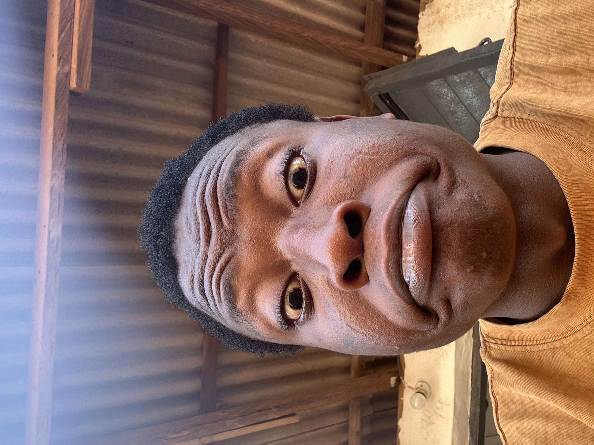 A close-up, portrait-style photograph captures a person with dark skin and short, dark, curly hair, seen from the chest up. The individual is positioned horizontally in the original image but appears to be looking upwards with an exaggerated, expressive facial gesture. Their eyes are wide open with golden-brown irises, and their forehead is visibly wrinkled, conveying a sense of surprise or intense concentration. The lips are pushed out into a distinct pout or exaggerated frown. The person is wearing a mustard yellow or light brown t-shirt, visible on the right side of the frame.
The background suggests an indoor or covered outdoor setting. Horizontally oriented corrugated metal sheeting is visible, likely forming a wall or ceiling. Wooden structural beams are interspersed throughout the background. In the lower right corner, an old-fashioned light bulb is mounted on a light-colored surface, which also shows faint, illegible markings or text. The lighting suggests natural daylight, illuminating the subject's face clearly. The overall mood conveyed by the subject's expression is one of humor or playful exaggeration.