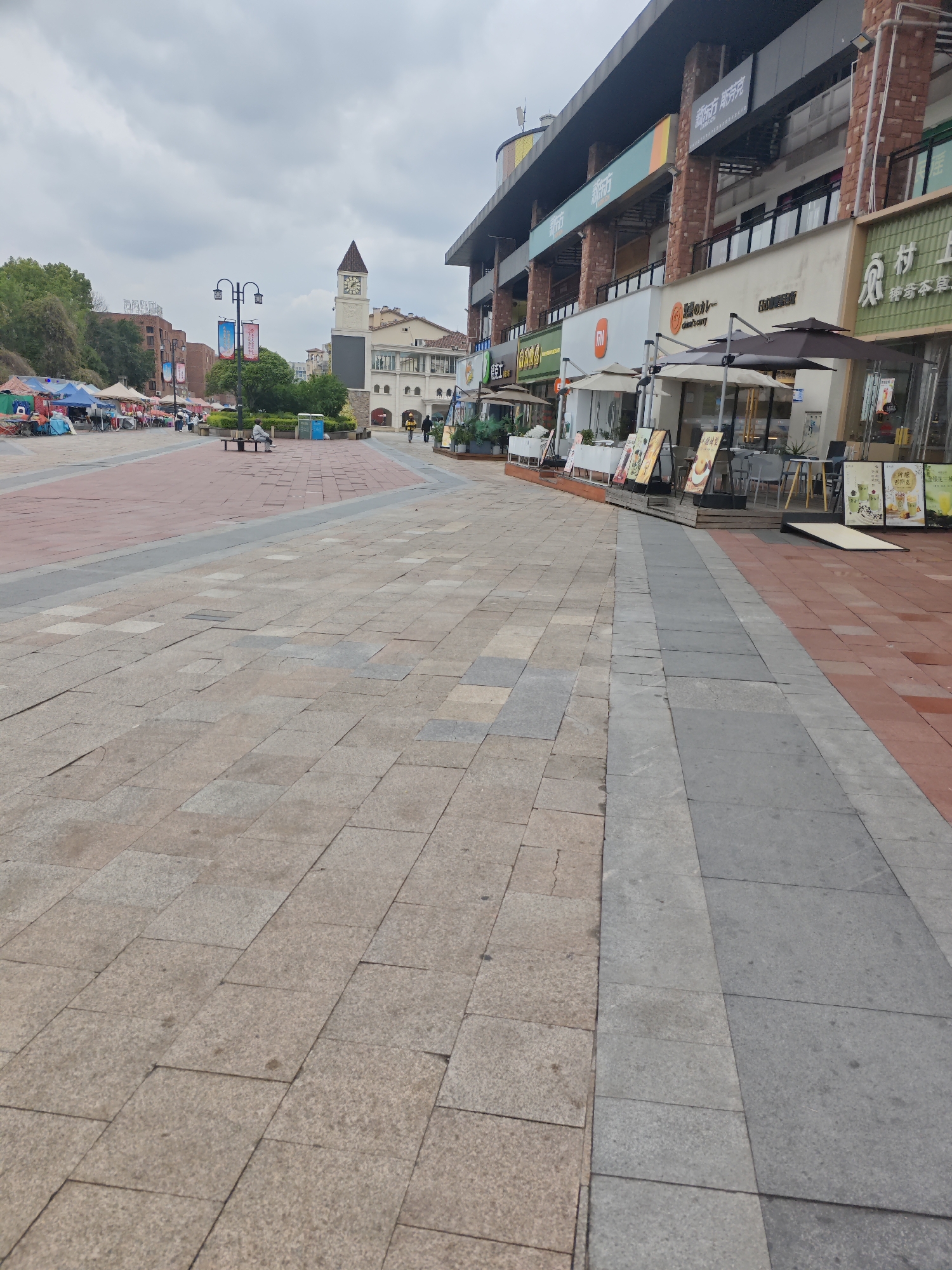 The image depicts a commercial plaza in the Chongqing High-tech Zone, China. The foreground shows a paved pedestrian area with a mix of tan, grey, and red bricks. To the left, under a cloudy sky, a line of vendors' tents and stalls can be seen. In the center distance, a clock tower stands near benches and people. To the right, a multi-story commercial building lines the plaza, with several businesses visible including a Xiaomi store, a curry restaurant, and other restaurants and shops with outdoor seating. Signage in Chinese characters is prominent.