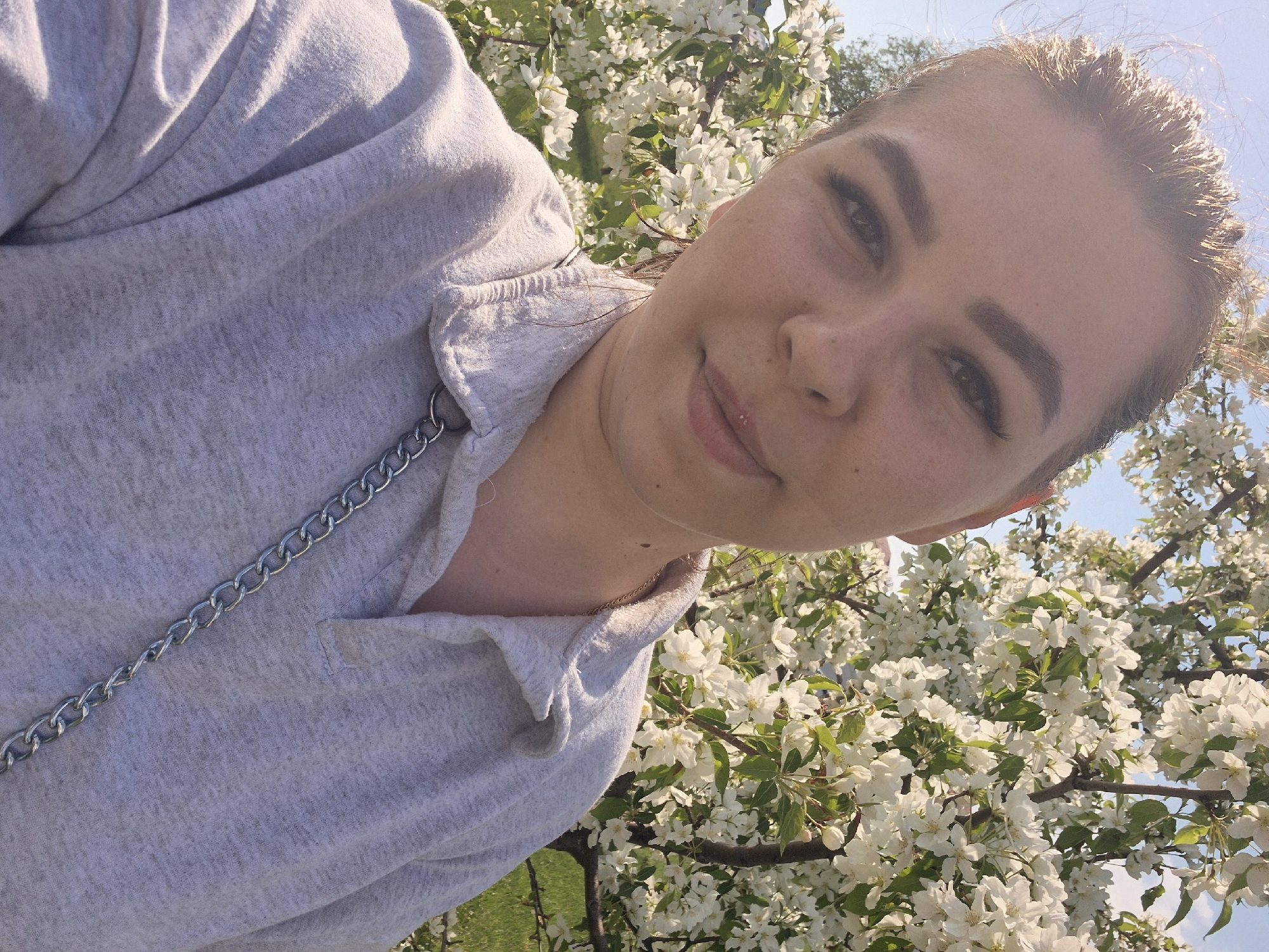 A woman in a gray sweatshirt stands in Tobolsk, Russia, surrounded by flowering trees with white blossoms. She is facing the camera with a slight smile. A silver chain is visible on her sweatshirt. The background includes a blue sky partially obscured by the branches and flowers.