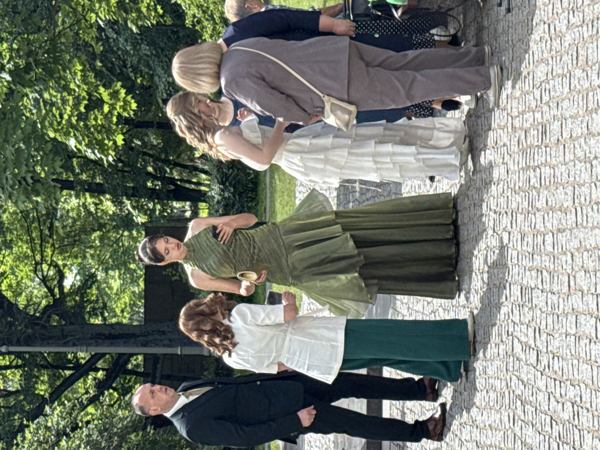 This media file captures a group of people in what appears to be a staged event or photoshoot. The subjects are positioned on a cobblestone path, with a backdrop of lush green trees. The lighting suggests it is daytime, with sunlight creating distinct shadows on the ground.
The individuals are dressed in a variety of attire, hinting at different roles or themes within the gathering. One person wears a dark suit, another a white blouse paired with green pants, and another a layered white dress. Two other individuals are also present, one in a green dress and another in a brown suit. The overall impression is one of elegance and perhaps a formal occasion.
There are no visible text elements in the image. The scene is set outdoors, likely in a park or garden area, given the surrounding foliage. The expressions on the faces of the people are neutral, making it difficult to ascertain specific emotions or the exact nature of the activity. The arrangement of the individuals and their clothing suggests a deliberate composition, possibly for a wedding, a historical reenactment, or an artistic project. The presence of cobblestone paving and the mature trees could indicate a historical or park-like setting.