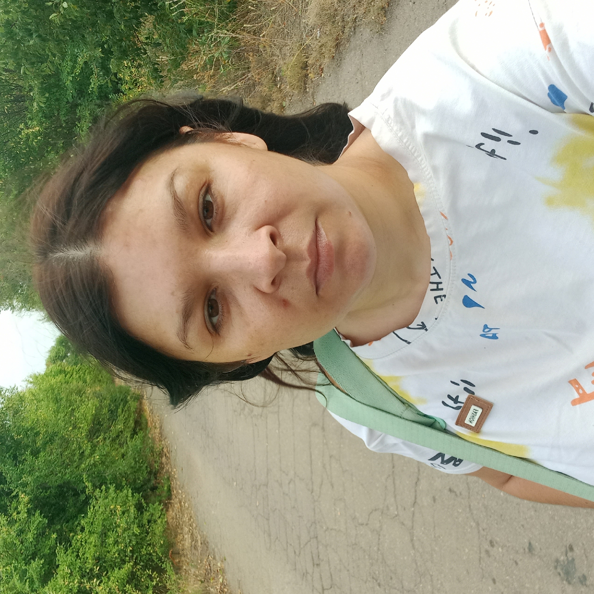 A medium shot selfie shows a woman standing on a paved road surrounded by dense green trees and dry grass. She has dark brown hair, pulled back in loose waves, and brown eyes. She is wearing a white t-shirt with a colorful paint splatter design and some handwritten text. A light green strap from a bag is visible over her right shoulder. A small rectangular tag with the word "FASHION" is attached to the t-shirt. The road appears weathered with visible cracks. The background shows a natural, unurbanized environment, consistent with a rural road.