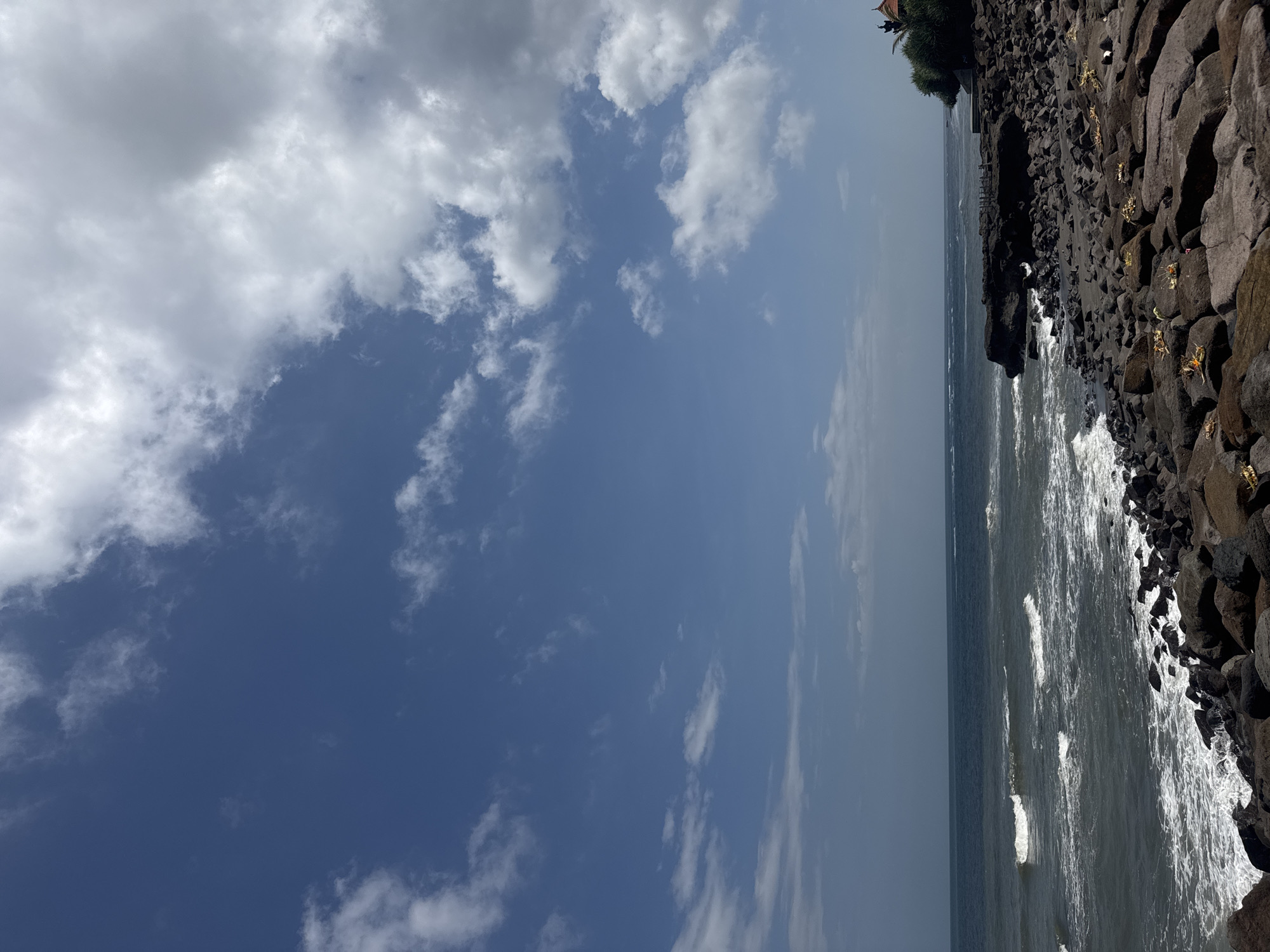 The image captures a coastline in Mengwi, Indonesia, featuring a rocky shore with black volcanic rocks and sparse green vegetation, interspersed with golden offerings. The ocean stretches out to the horizon, with visible waves crashing against the shore. The sky is blue with scattered white clouds.