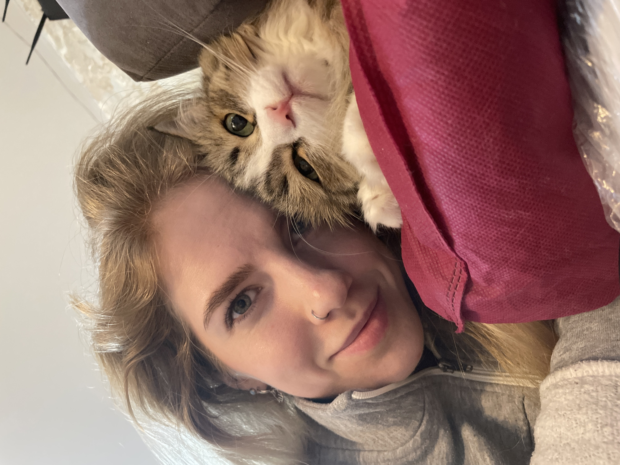A selfie-style photograph shows a fair-skinned woman with blonde hair and blue eyes looking directly at the camera. She has a nose ring and is wearing a gray hooded sweatshirt. Her head is tilted to the side, and a cat with green eyes, brown and white fur, and long whiskers is positioned above her head, partially obscured by what appears to be a burgundy shopping bag and a gray cushion. The background is a white wall. There is no information to suggest the location is Izhevsk, Russia.