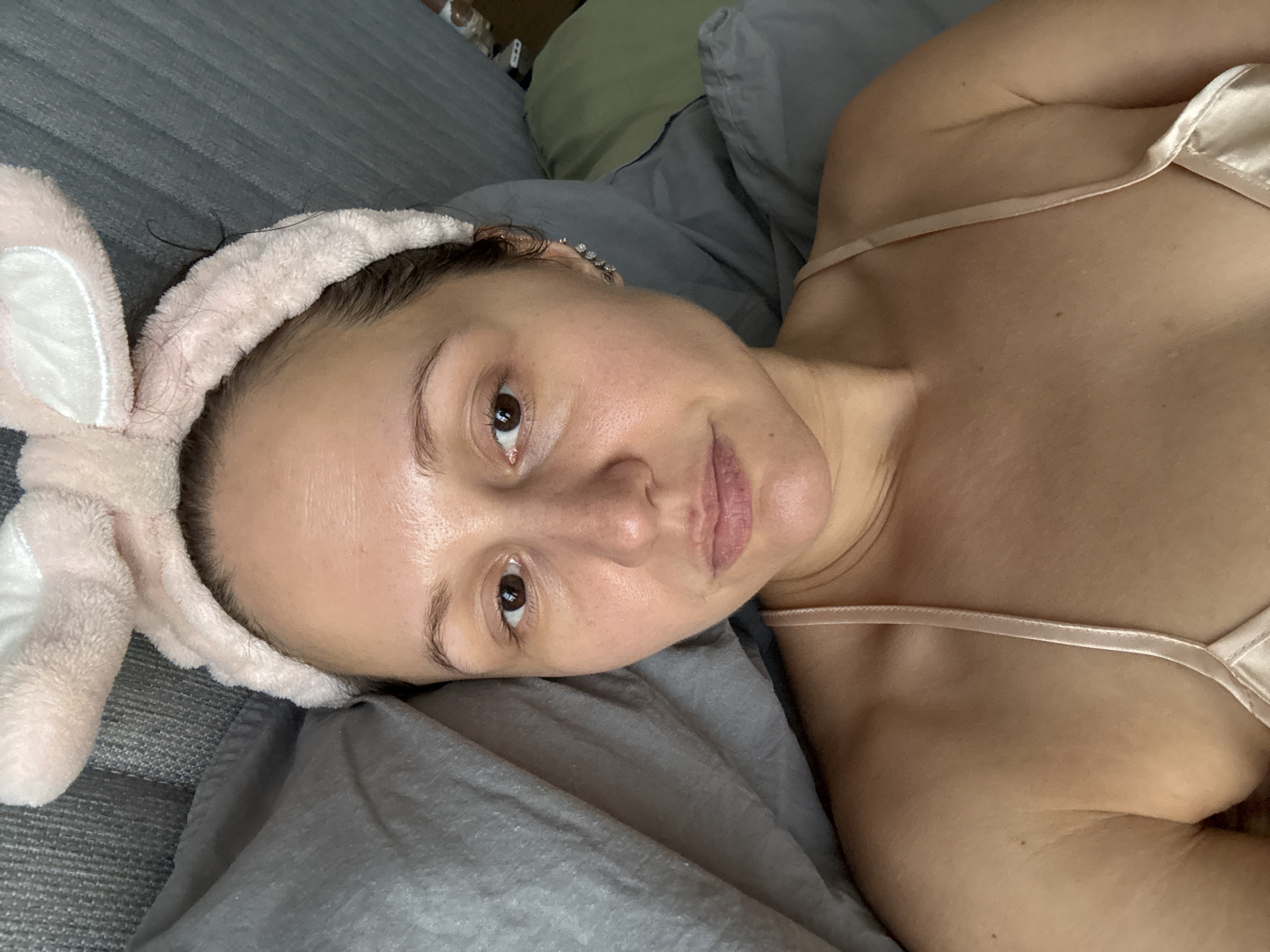 A woman is lying on a gray bed, wearing a light pink satin camisole and a pink bunny ear headband. Her head is resting on a gray pillow. Her facial features include brown eyes and lips, and fair skin. No elements in the image suggest a location in Dubai, United Arab Emirates. The setting appears to be a bedroom.