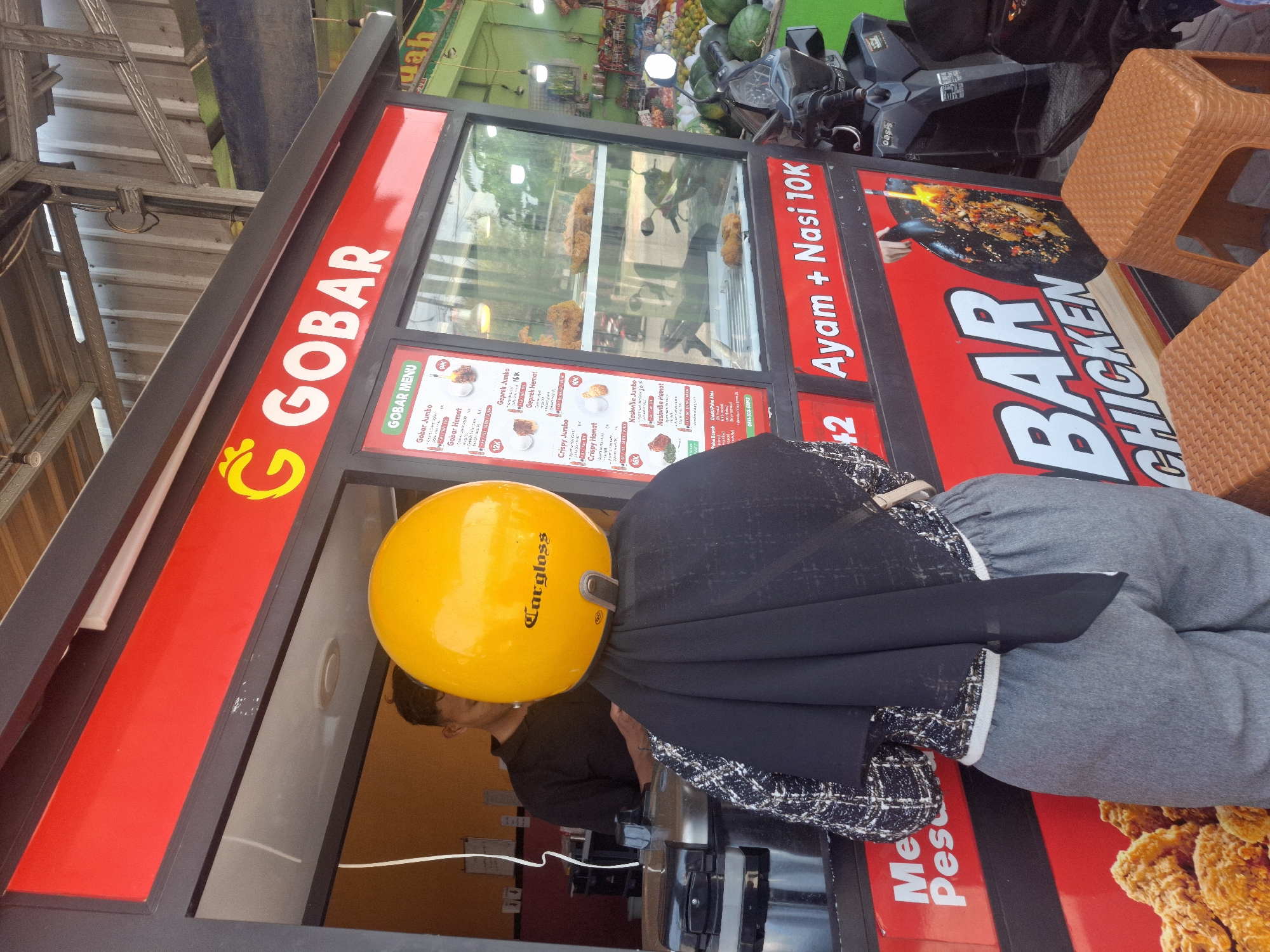 In Sumberwudi, Indonesia, a person wearing a yellow helmet and a black and white patterned jacket stands in front of a food stall called "GOBAR". The stall has a red and black exterior with menus displayed in a glass window, showcasing fried chicken items. A man can be seen inside the stall. The person in the foreground is facing away from the camera, with their back to the viewer. In the background, a motorcycle is parked next to the stall, and a market with fruits and vegetables is visible. The overall scene depicts a street food vendor operation.