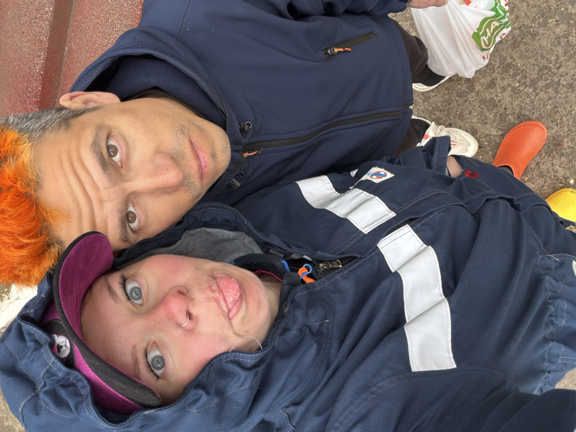 Two individuals are lying on a concrete surface. One individual has orange hair and is wearing a blue jacket. The second individual, located below and slightly to the side, is wearing a pink and black baseball cap and a blue jumpsuit with reflective stripes. The second individual's tongue is protruding from their mouth. The background includes an orange boot, a yellow object, and a white plastic bag with green writing. The location is the city of Chaikovsky, Russia.