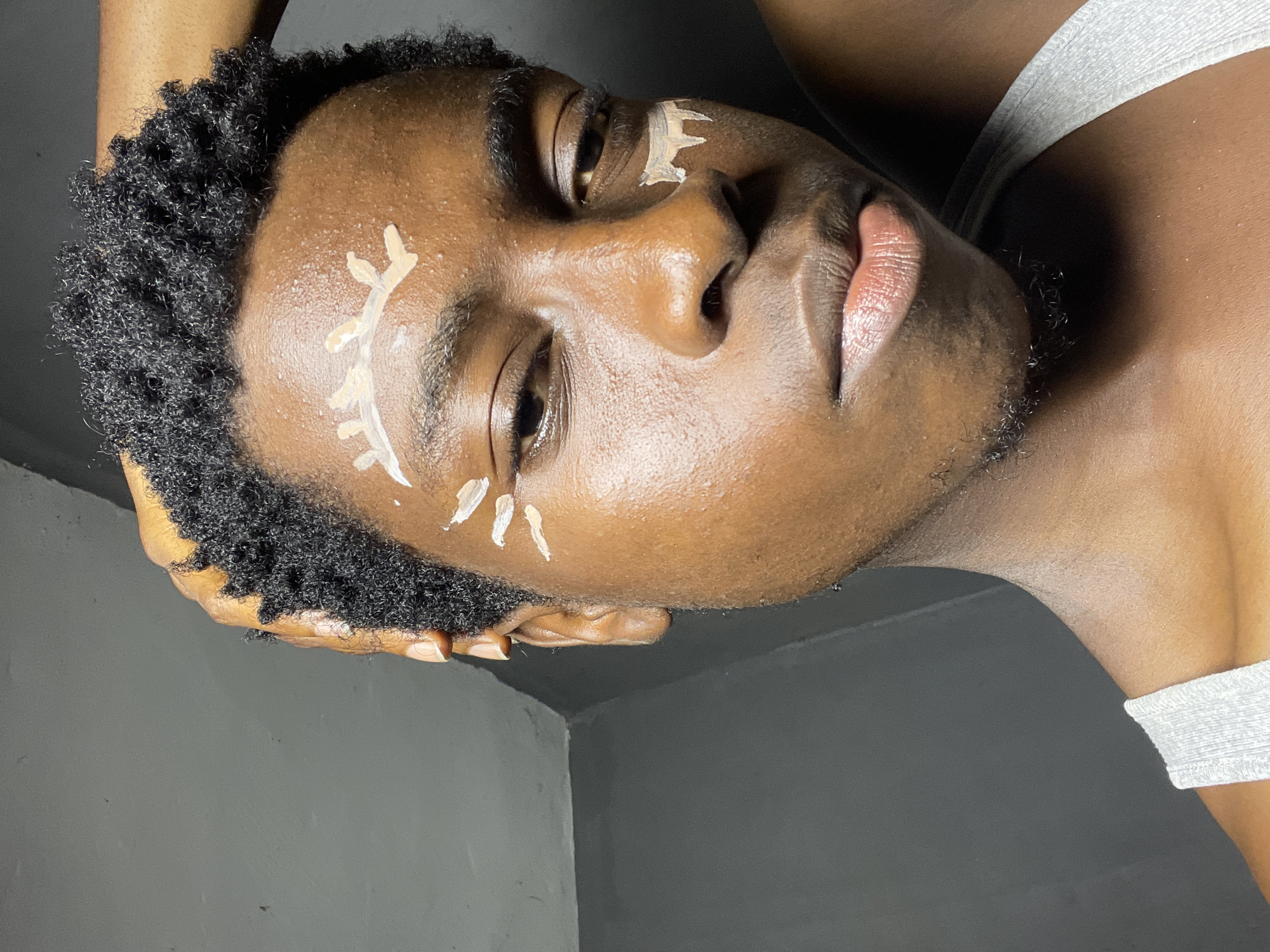 The image is a close-up, waist-up portrait of a young Black male with dark, curly hair, touching his head. He is wearing a grey tank top. There are several streaks of a light-colored substance, possibly makeup or paint, applied to his forehead and cheek. The background is a plain, dark wall with a visible seam or corner. The lighting appears to be artificial and creates shadows on his face and the background. The subject's expression is neutral to slightly melancholic, with his gaze directed towards the camera. The image does not contain any information that would allow for the determination of its location within Nkpor, Nigeria.