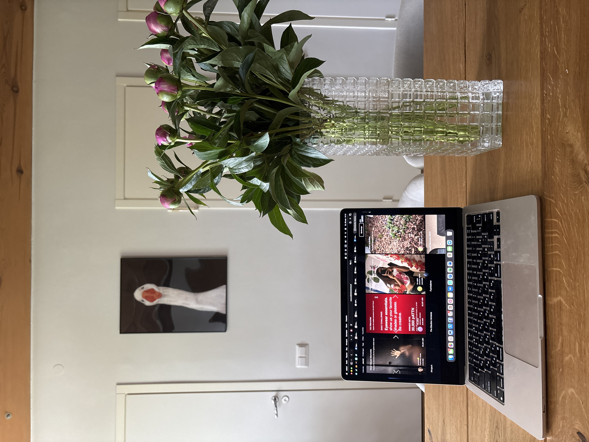 On a light brown wooden table, a silver laptop is open, displaying a screen filled with various images and text in a social media interface. Next to the laptop, a clear glass vase holds a bouquet of peonies, with green leaves and buds. The background consists of white walls with a white door and a picture of a white goose on the wall. The image was not taken in Chaikovsky, Russia.