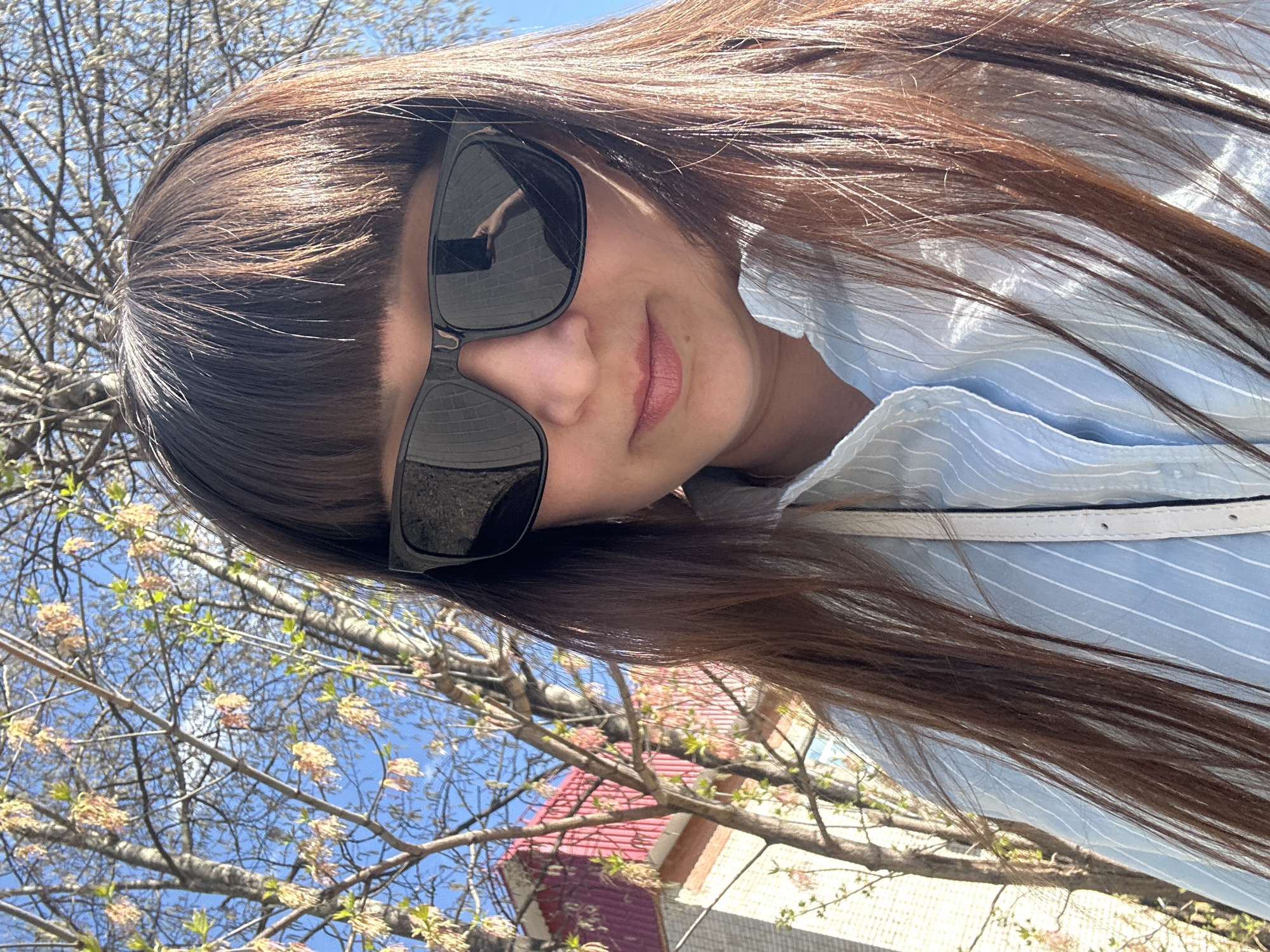 A woman with long, dark hair and sunglasses is the primary subject of the image. She is wearing a light blue striped shirt with a white strap across her shoulder. The background features branches of a tree with light-colored flowers against a blue sky. A building with a red roof is also visible. The image appears to be taken in an outdoor setting, possibly a park or street. Based on the prompt the location is Chaikovsky, Russia.