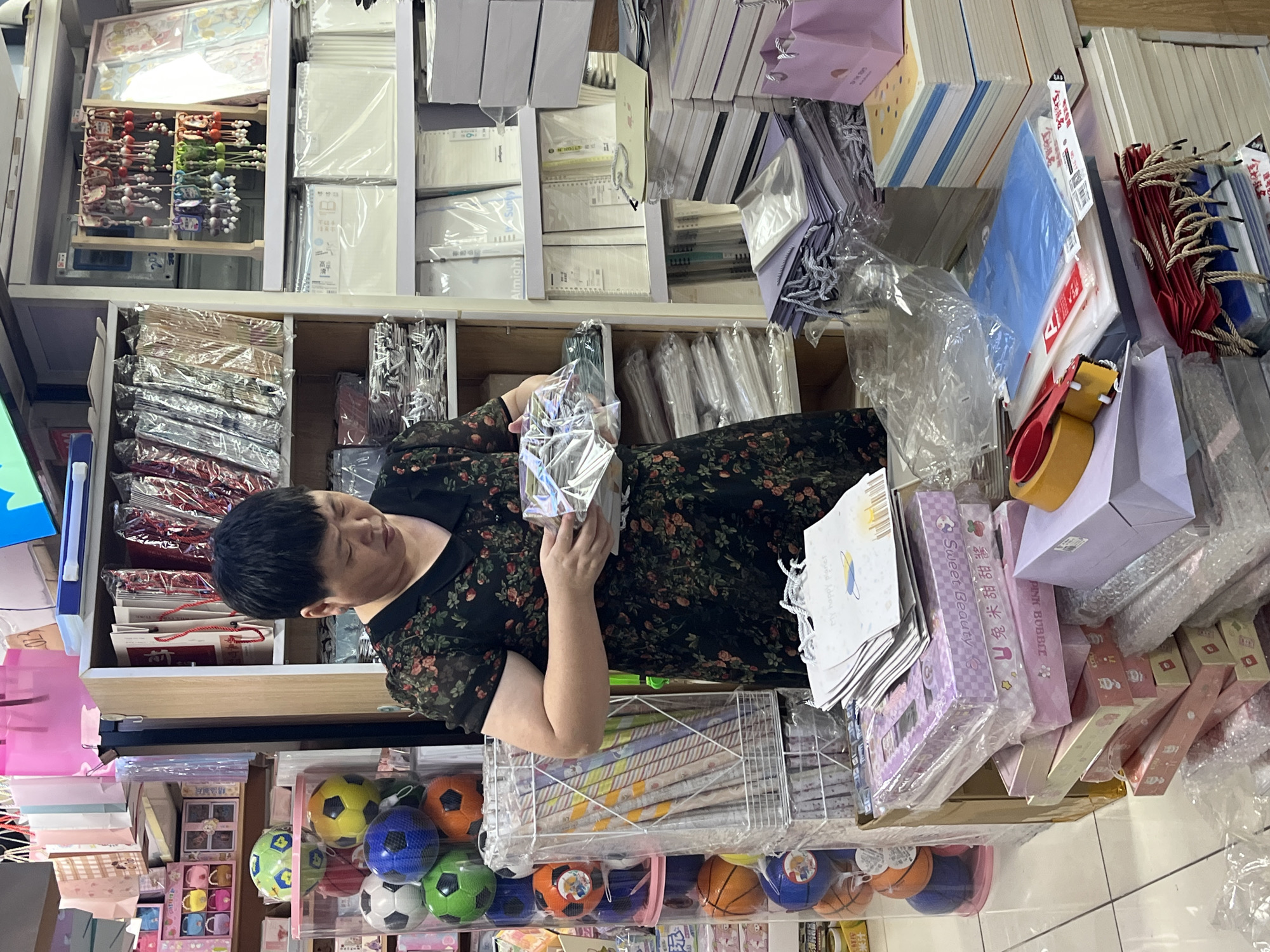 A medium shot captures a woman in a floral dress standing in a shop in Liwan District, China, surrounded by merchandise. She is holding several items wrapped in clear plastic. The shop is filled with shelves stocked with various goods, including colorful toy balls, stationery supplies like notebooks and paper, and gift bags. In the foreground, stacks of boxes and paper products are visible. The overall environment suggests a retail setting, likely a stationery or gift shop.