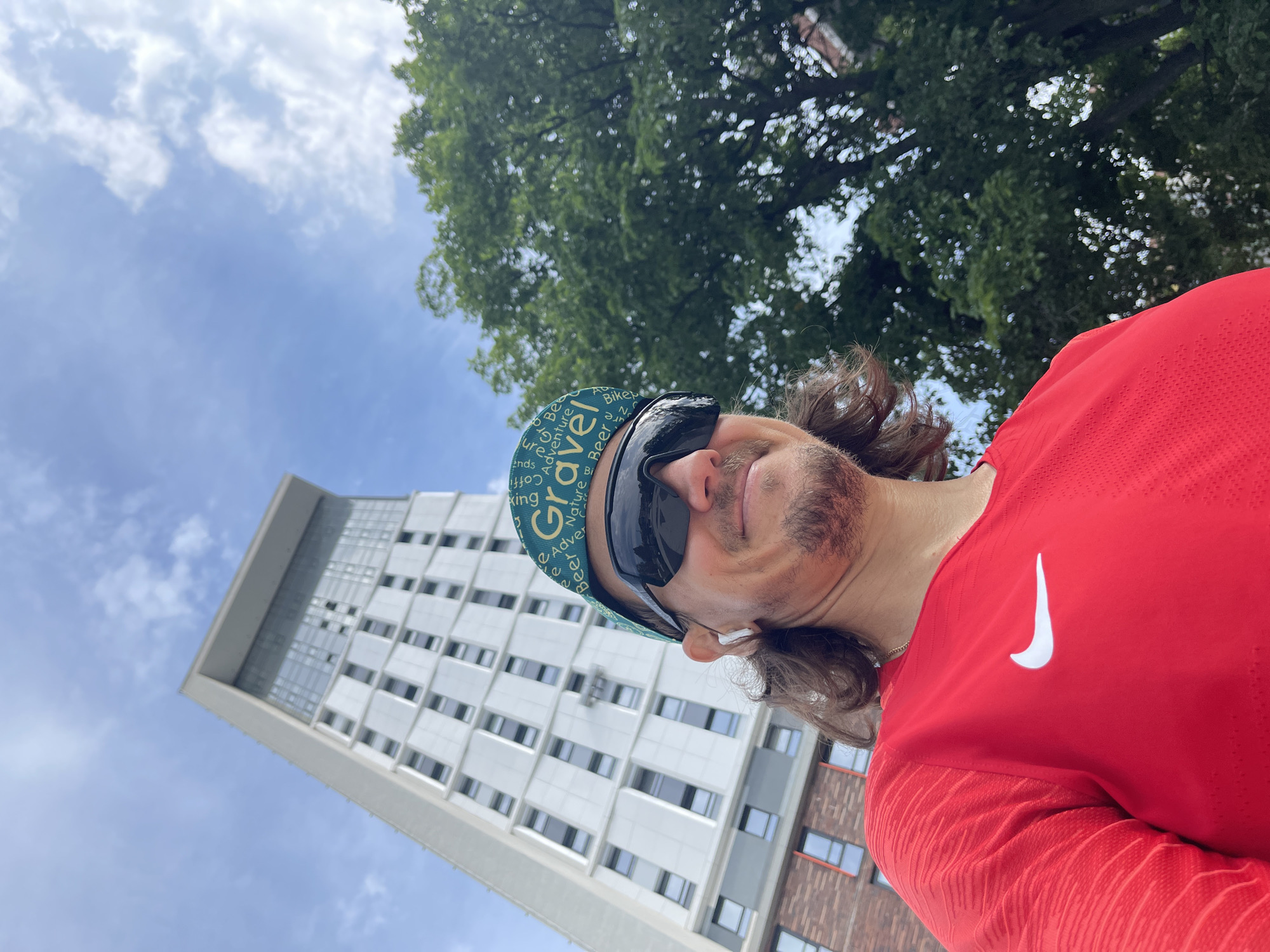 A tilted selfie photograph captures a man outdoors on a bright day. He is seen from the chest up, positioned on his side. He wears a vibrant red Nike t-shirt, clearly displaying the white Nike swoosh logo on his left chest. On his head, he sports a green cycling-style cap with various white words printed on it, including "Gravel," "Adventure," "Coffee," "Beer," "Biking," "Nature," "Rides," and "Friends." Dark, wraparound sunglasses obscure his eyes, and a white earbud is visible in his right ear. He has brown, curly hair and some facial stubble, and a slight smile or smirk plays on his lips.
The background is divided into two main elements under a clear blue sky with some wispy white clouds. On the left, a tall, modern, light-colored building with multiple floors and rectangular windows stretches upward. On the right, a large, leafy green tree fills the upper portion of the frame, providing a natural contrast to the urban structure. The bright lighting suggests it is daytime. The overall impression is of an active individual capturing a moment in an urban or suburban setting.