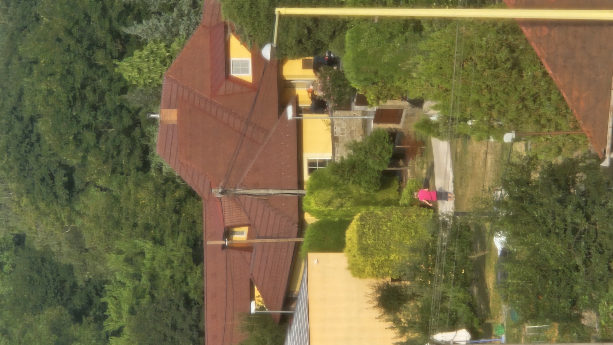 The image depicts a residential area with a large house featuring a red tiled roof and yellow walls, surrounded by dense green trees and manicured hedges. A person wearing a pink shirt and dark shorts is walking on a paved path in the foreground. The scene appears to be a suburban or rural setting, possibly in the city of Prešov, Slovakia, given the architectural style and the lush greenery. There are streetlights visible, and power lines run across the scene, suggesting a developed area.