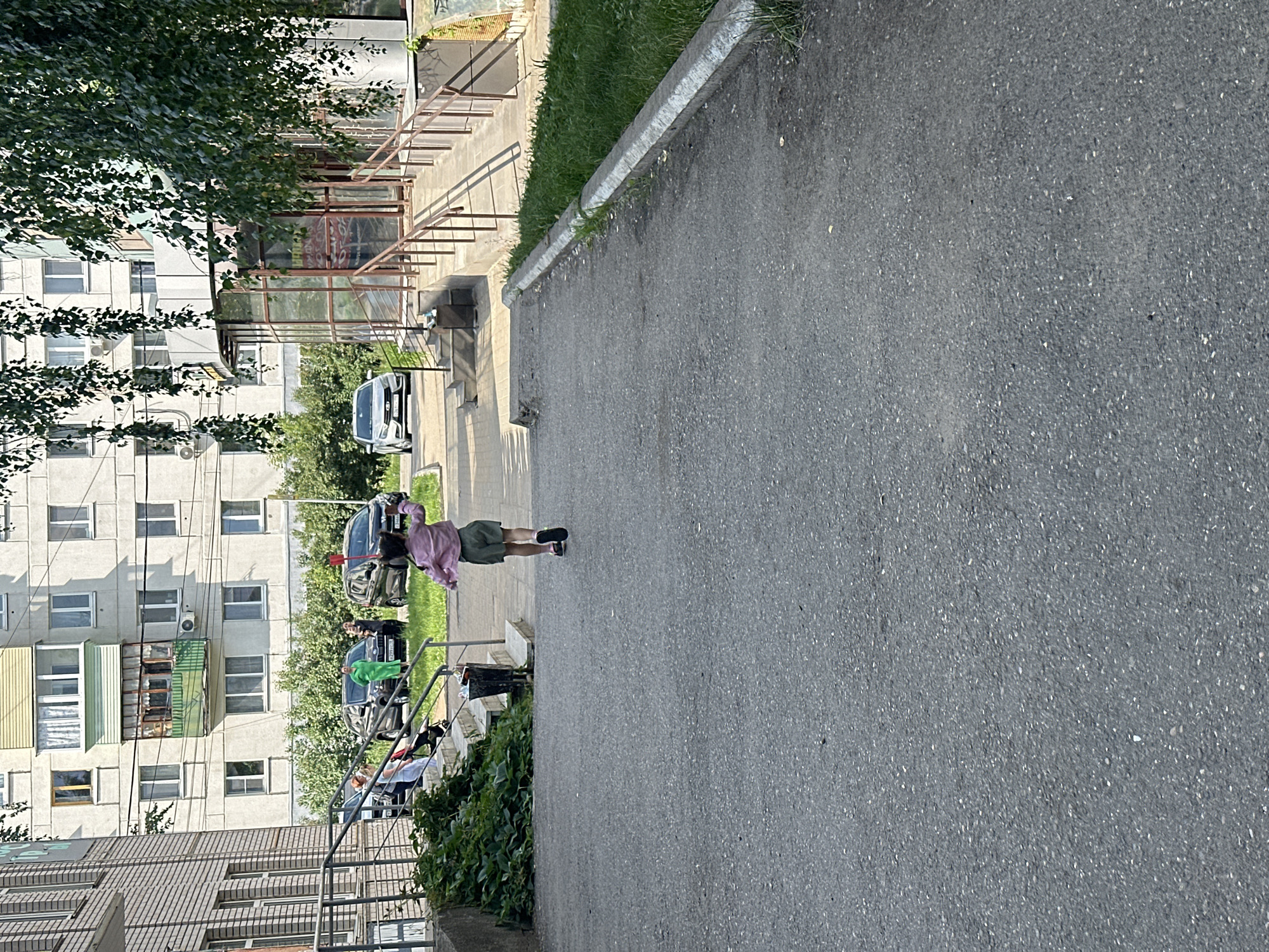 A young child, viewed from behind, runs away from the camera down an asphalt pathway. The child wears a pink, possibly satin, shirt and olive green shorts. The pathway is bordered on the right by a strip of green grass and then a concrete curb. In the background, a multi-story apartment building with many windows and balconies rises above a row of trees and bushes. To the left, a staircase leads up to a building entrance with a metal railing. Several parked cars are visible near the building entrance, and a few people are gathered on the staircase. A trash can is situated at the base of the stairs. The lighting suggests a sunny day.