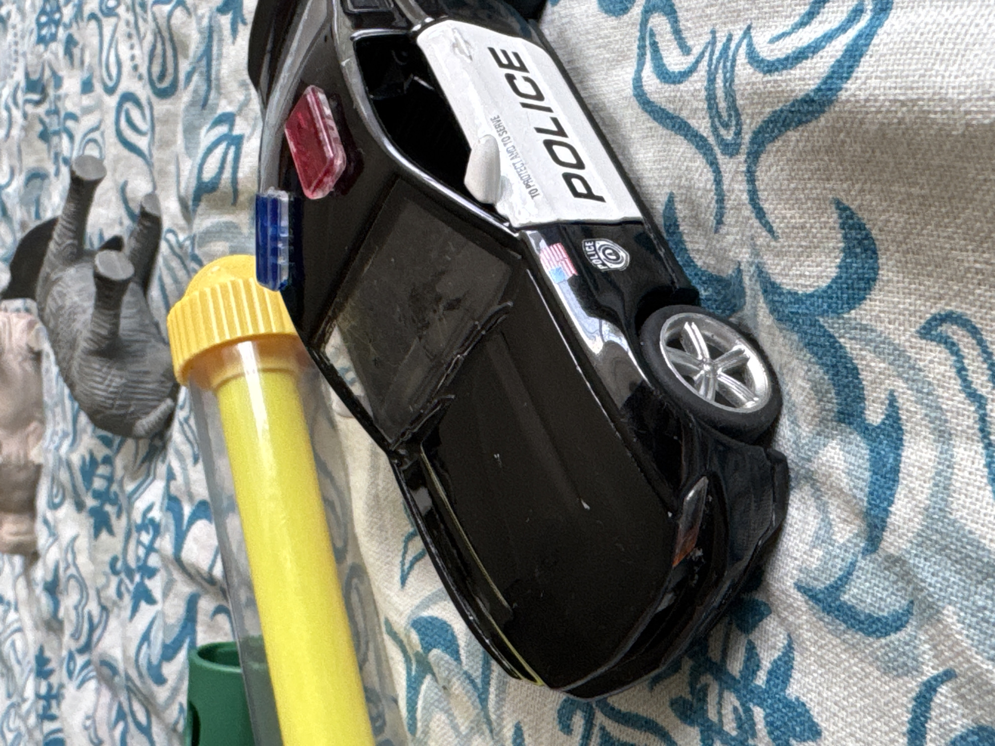 The image shows a close-up of a black toy police car, possibly a Ford Mustang, with "POLICE" written in white on the side. It has a blue and red light bar on its roof. To the left of the car is a yellow cylindrical object with a yellow ribbed end, and a green object partially visible. The background is a patterned fabric, likely a blanket or bedsheet, with a blue and white design. There is no indication of the city of Яромінскі сельскі Савет, Belarus, or any people or actions taking place.