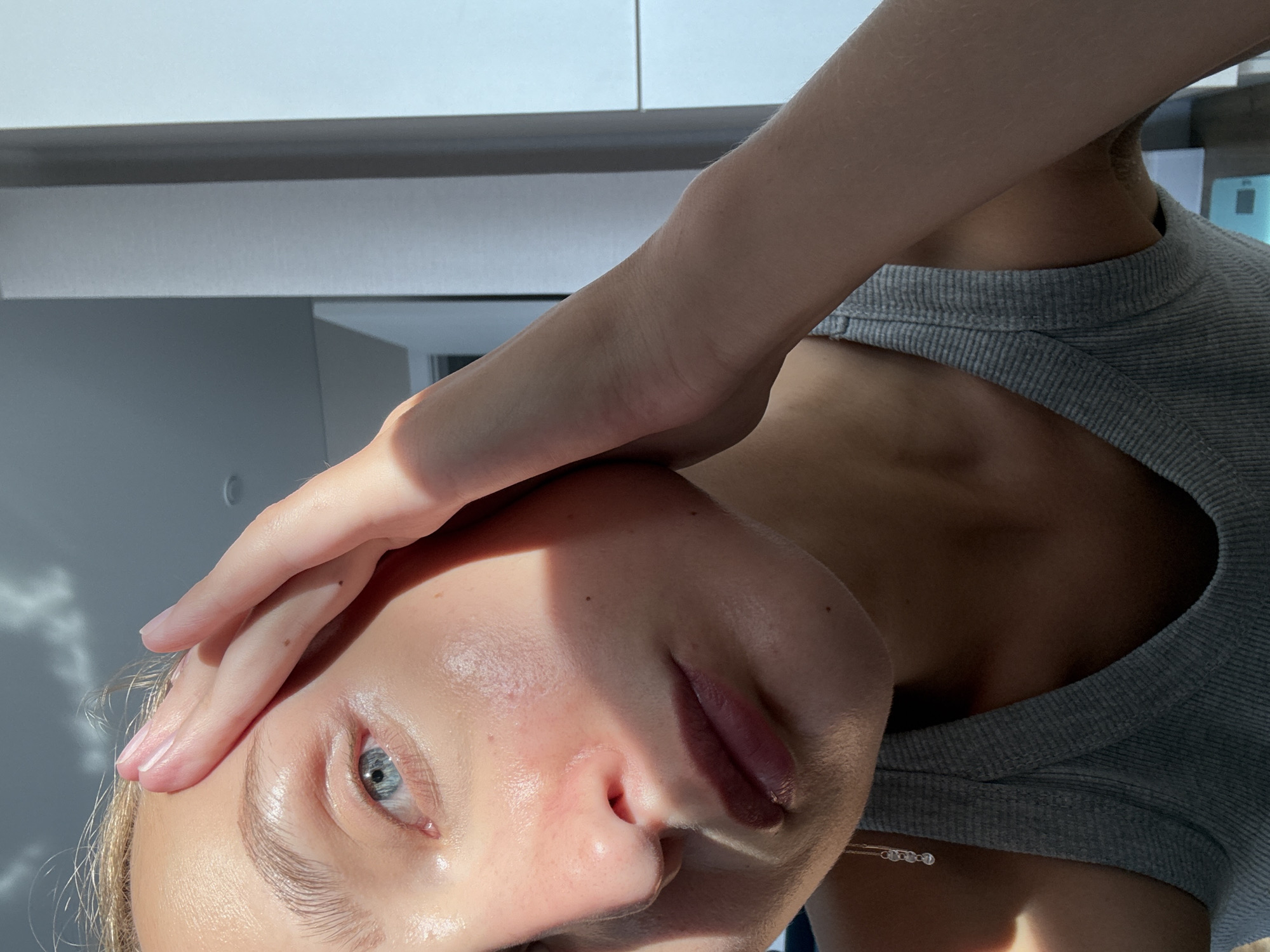 A close-up, low-angle indoor shot features the face of a young woman positioned on the left side of the frame. She is wearing a grey tank top and has her left hand resting on her forehead, obscuring part of her face. Her blue eyes are visible, and her lips are slightly parted. Sunlight casts shadows across her face and skin, highlighting a dewy complexion. The background is out of focus, showing a mostly white wall and a sliver of what appears to be a door or another architectural element. The overall lighting suggests a bright, possibly daytime setting. There is no indication of the city of Подшивалово, Russia, or any other specific location.
