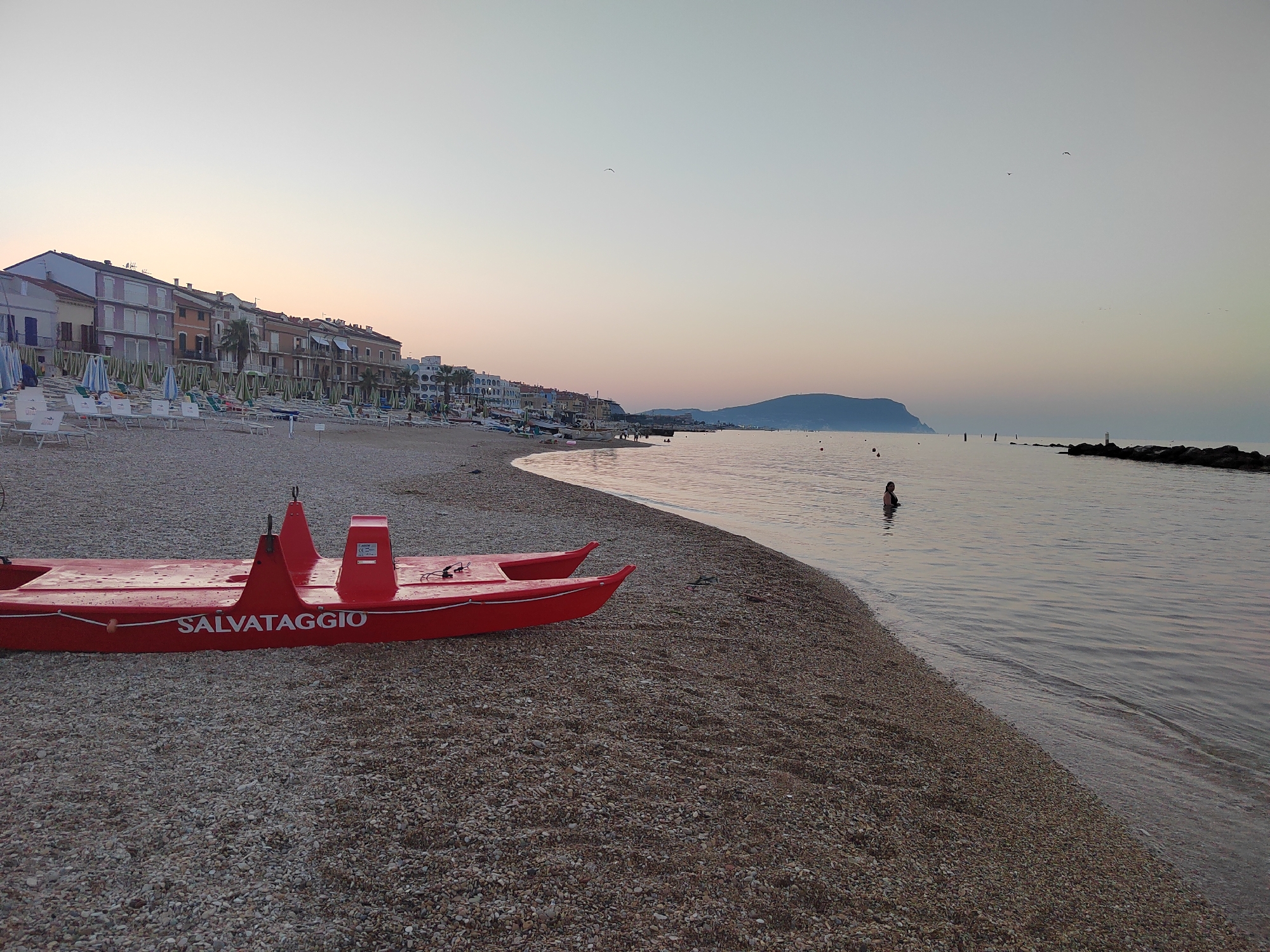 This image depicts a serene beach scene in Porto Recanati, Italy, at what appears to be either sunrise or sunset. In the foreground, a bright red, inflatable rescue boat with "SALVATAGGIO" printed on its side rests on a pebble beach. The boat is positioned to the left of the frame, angled towards the water. The beach is covered in smooth, multi-colored pebbles and extends towards the right edge of the image.
To the left, lining the shore, are numerous rows of closed beach umbrellas and lounge chairs, suggesting a resort or public beach area that is not currently in active use. Behind the beach chairs and umbrellas, a strip of buildings, likely hotels or residences, forms a backdrop. These buildings have a typical Mediterranean architectural style with multiple stories and balconies.
The water of the Adriatic Sea is calm and reflects the soft hues of the sky. In the mid-ground, a solitary person is visible standing in the water, their back to the camera. Further out in the sea, a distant headland or island is visible, silhouetted against the pale sky. A few small buoys and a breakwater made of rocks are also present in the water. The overall atmosphere is tranquil and peaceful, characteristic of the early or late hours of the day.