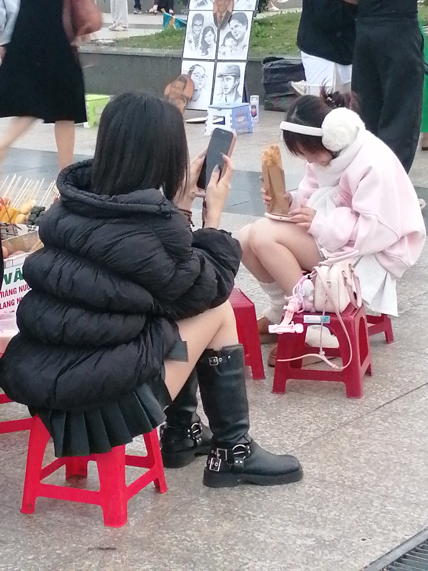 The image depicts two young females sitting on red plastic stools in an outdoor urban setting, likely in Da Lat, Vietnam, given the context of similar street scenes in that city. The foreground features a young female on the left wearing a voluminous black puffer jacket and a pleated black skirt. She is sitting on a bright red stool and is captured from the mid-torso down, with her black knee-high boots with buckles visible. Her hands are obscured by her jacket, but she appears to be holding a dark smartphone.
To her right, a second young female is seated on another red stool, facing slightly away from the viewer. She is wearing a light pink hooded jacket over a white skirt and white leg warmers. Her attention is focused on a brown paper-wrapped item she is holding and looking at. Beside her, on the stool, is a small light pink handbag with various charms attached.
The background is blurred, but it suggests an area with other people, possibly vendors, and what appears to be a display of framed sketches or drawings. The ground surface is paved with a speckled material. The overall scene suggests a casual, possibly public, gathering place where food or goods are sold.