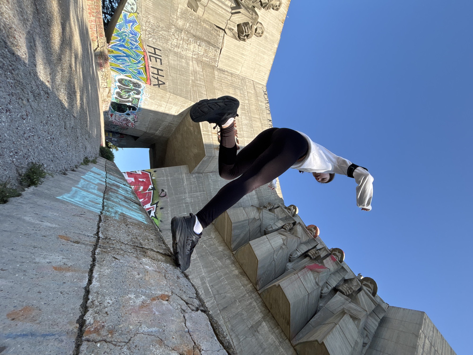 A dynamic, low-angle photograph features a person performing an inverted, acrobatic pose against a backdrop of massive concrete structures. The individual, dressed in black leggings, a white long-sleeved top, and black athletic shoes, has their right foot braced against a textured concrete ledge, while their body arches upside down towards the clear blue sky.
The scene is set at a brutalist-style concrete monument, identifiable as the Monument to the Bulgarian-Soviet Friendship in Varna, Bulgaria. The weathered concrete surfaces display extensive graffiti, with the distinct black block letters "HE HA" visible on a vertical section. Other colorful and less legible tags are present on the walls. The bright, clear daylight suggests a sunny day, with pronounced shadows on the concrete, indicating either morning or afternoon. The overall impression is one of urban exploration and athletic endeavor within an imposing architectural landscape.